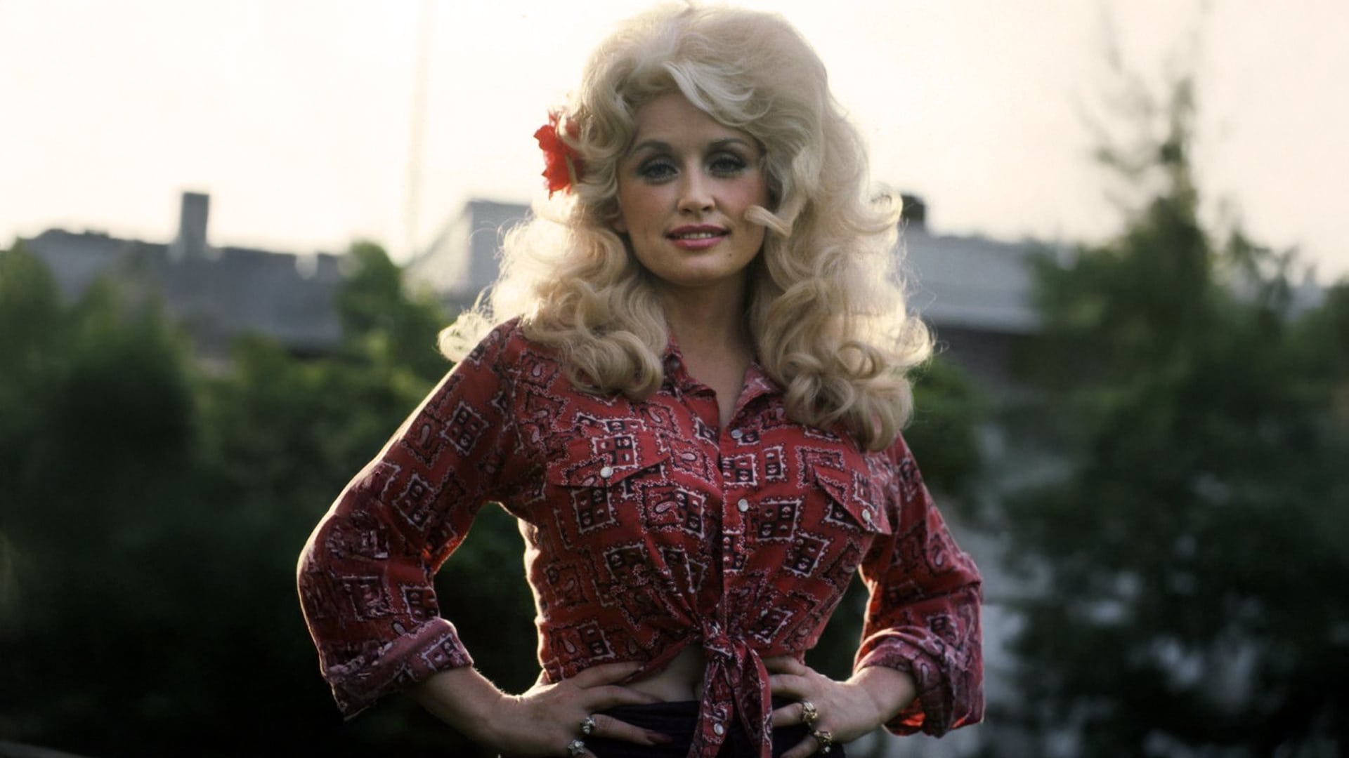 Dolly Parton stands confidently outdoors in a red patterned blouse, hands on her hips, as the article revisits the mystery of "Jolene" and imagines what the famously beautiful woman might have looked like.