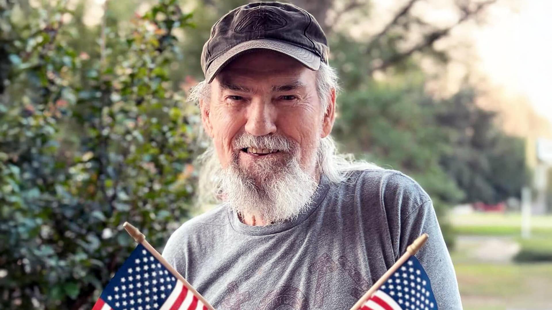 Uncle Si Robertson from Duck Dynasty smiles while holding two American flags, showing the quirky spirit behind the ten surprising facts about his life.