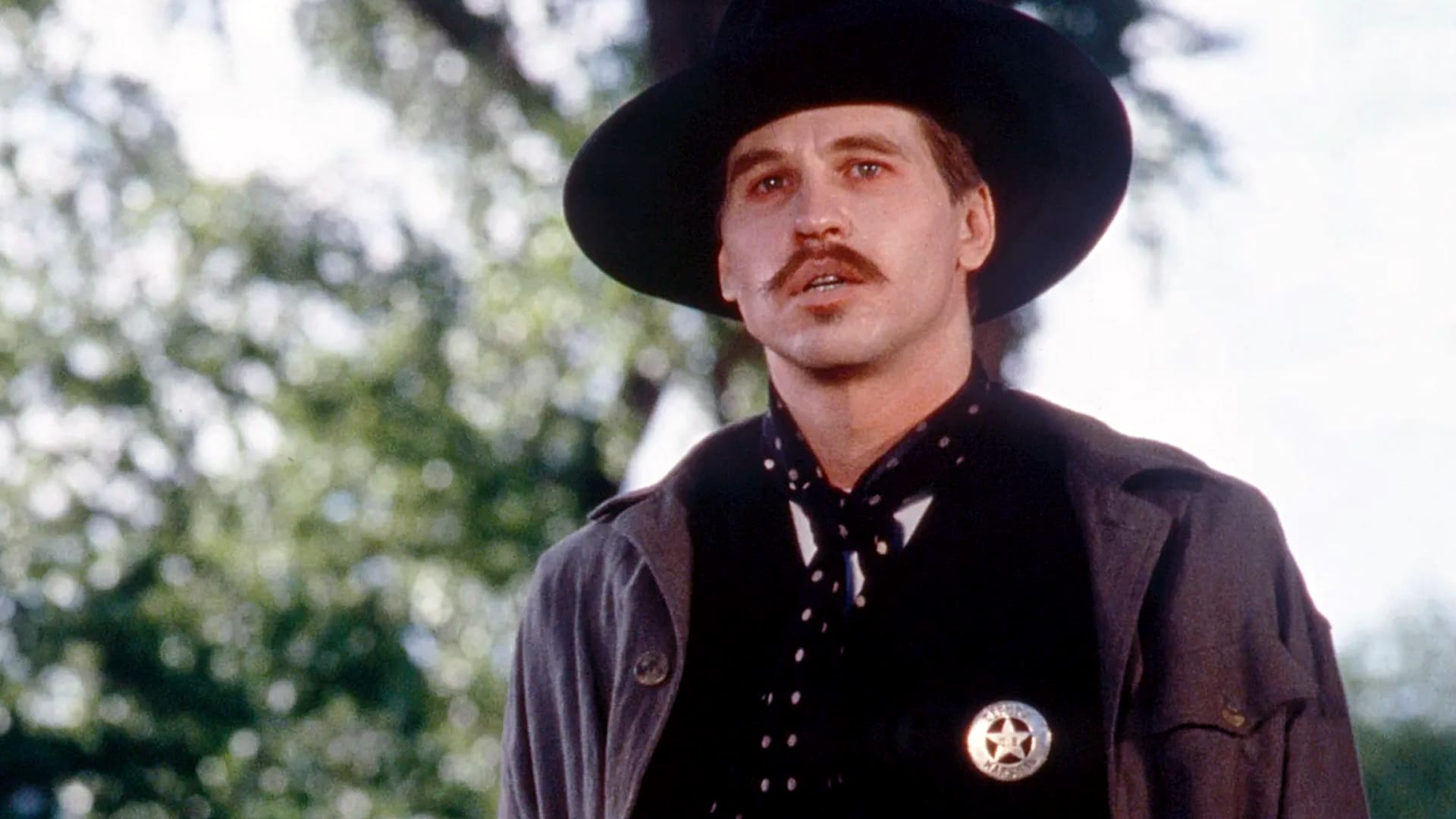 Val Kilmer in full Doc Holliday costume from Tombstone, wearing a black hat and vest, with a solemn expression and signature mustache.