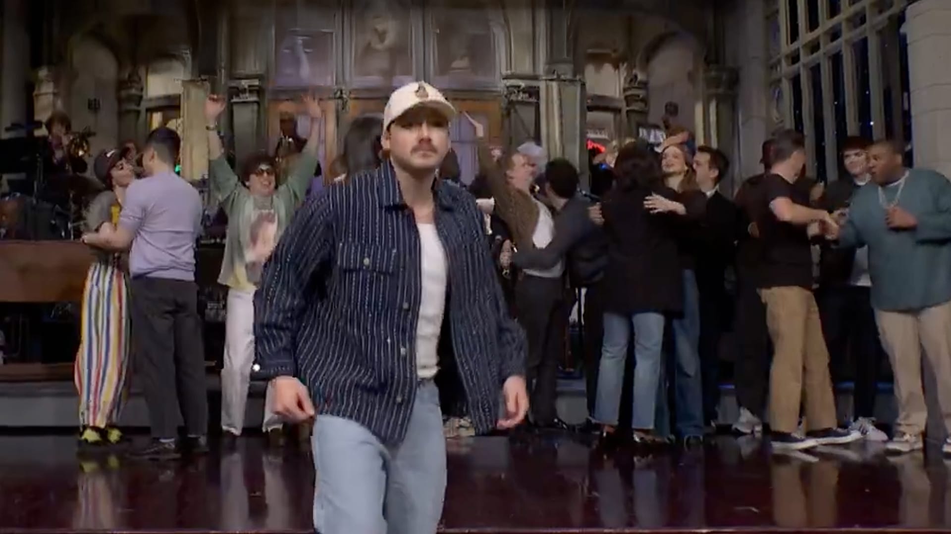 Morgan Wallen stands at the edge of the SNL stage in a dimly lit studio, leaning toward the host with a quiet expression just before walking off.