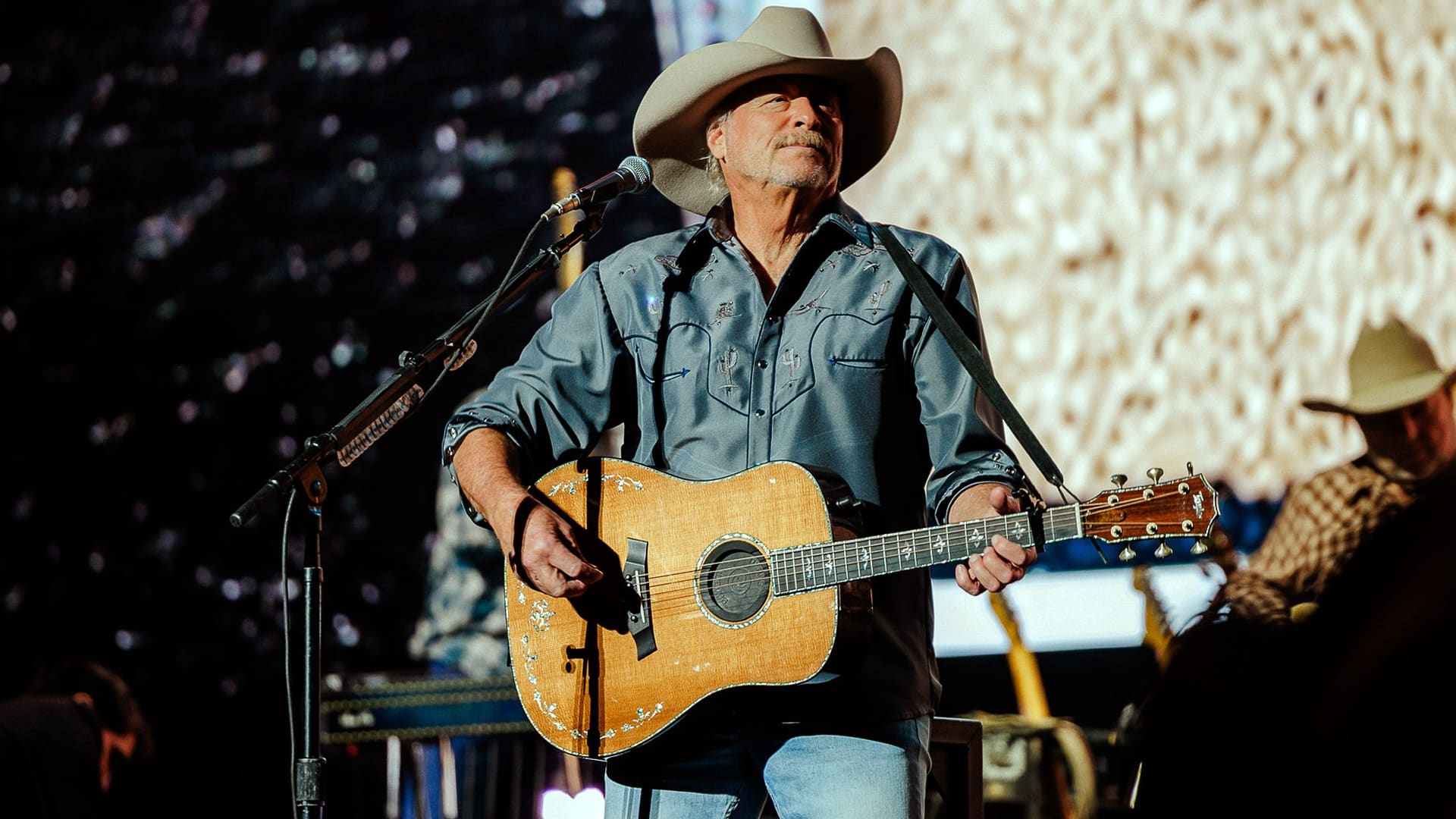 Alan Jackson performing on stage in a cowboy hat and guitar, gesturing toward the crowd during his farewell show at the Two Step Inn festival in Texas.