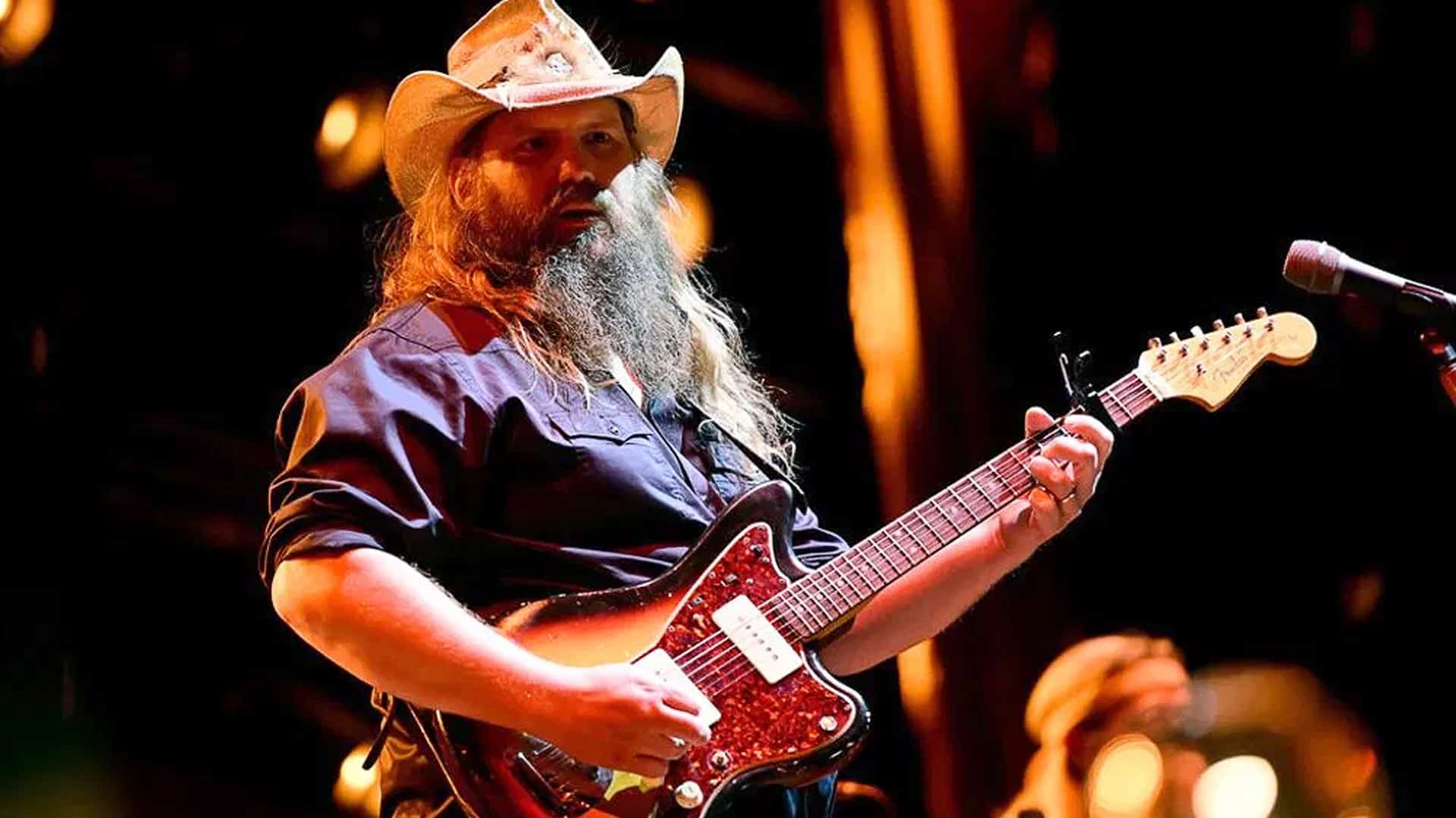 Chris Stapleton stands on stage wearing a cowboy hat and dark shirt, playing electric guitar with intense focus during a live performance.
