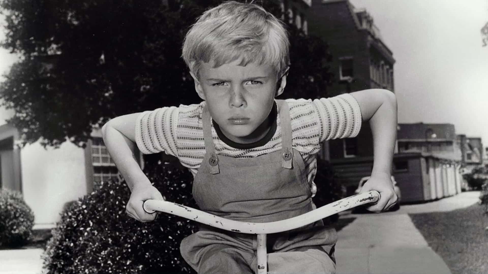 Black-and-white photo of a young boy with a determined expression riding a bike, portraying the classic TV character Dennis the Menace.