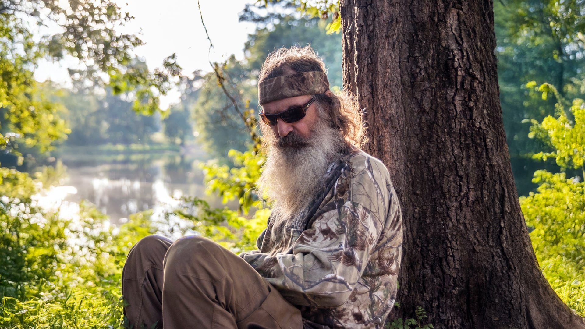 Phil Robertson from Duck Dynasty sits quietly under a tree in full camouflage, wearing sunglasses and a headband, deep in thought beside a peaceful wooded lake.