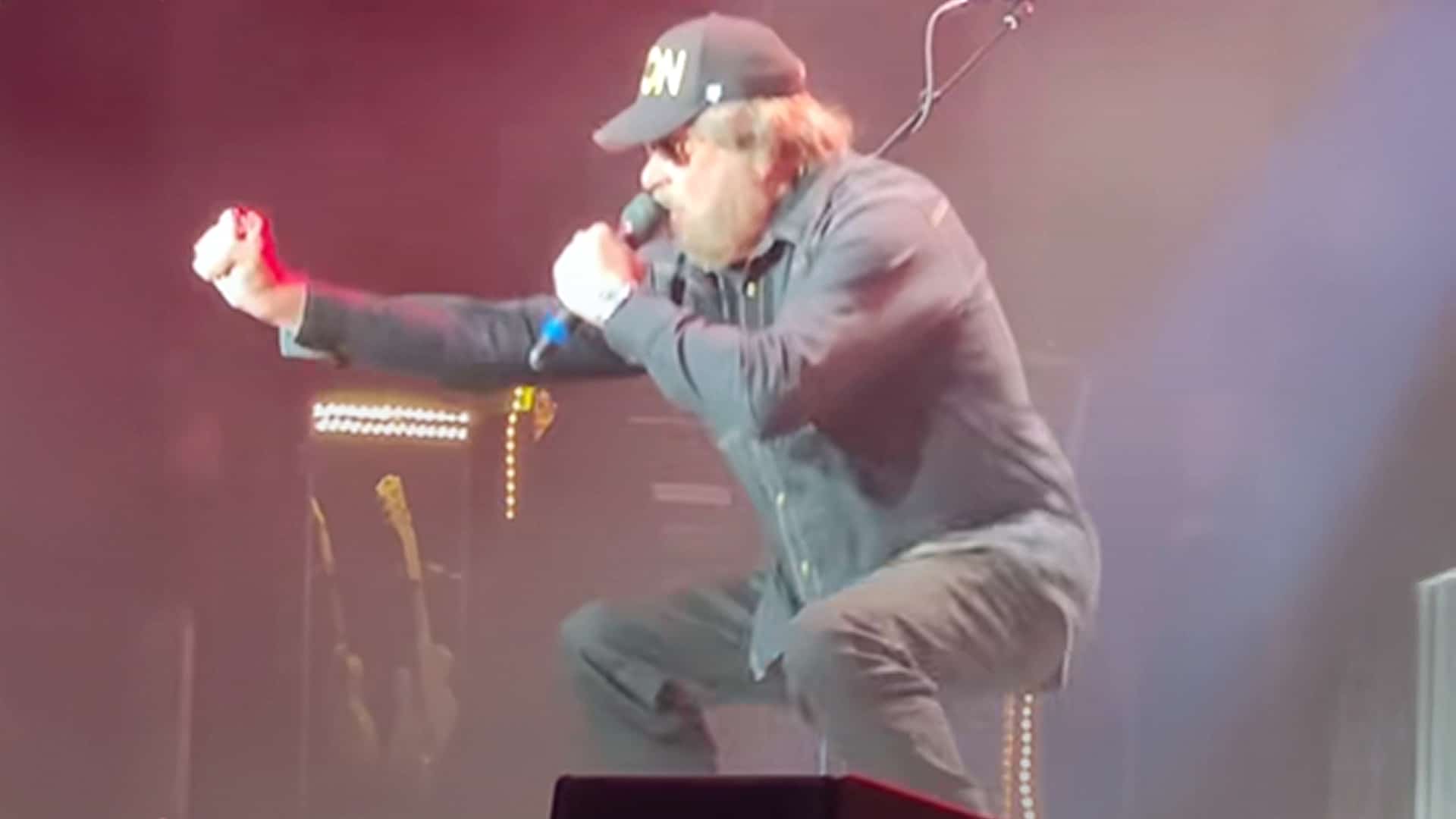 Hank Williams Jr. mid-performance, squatting low on stage with a mic in hand, wearing sunglasses and a black cap, showing off unexpected agility at age 75.