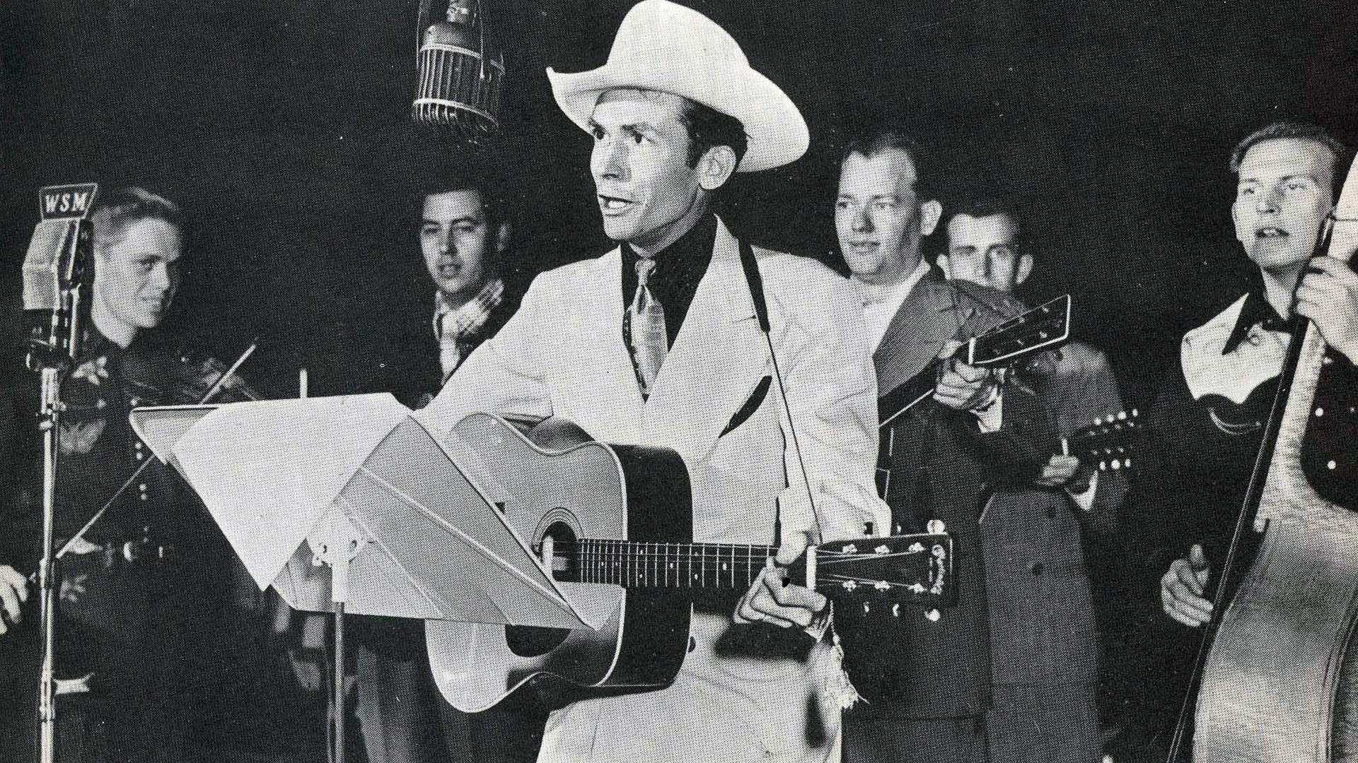 Hank Williams performs in a classic cowboy hat and white suit, surrounded by his band