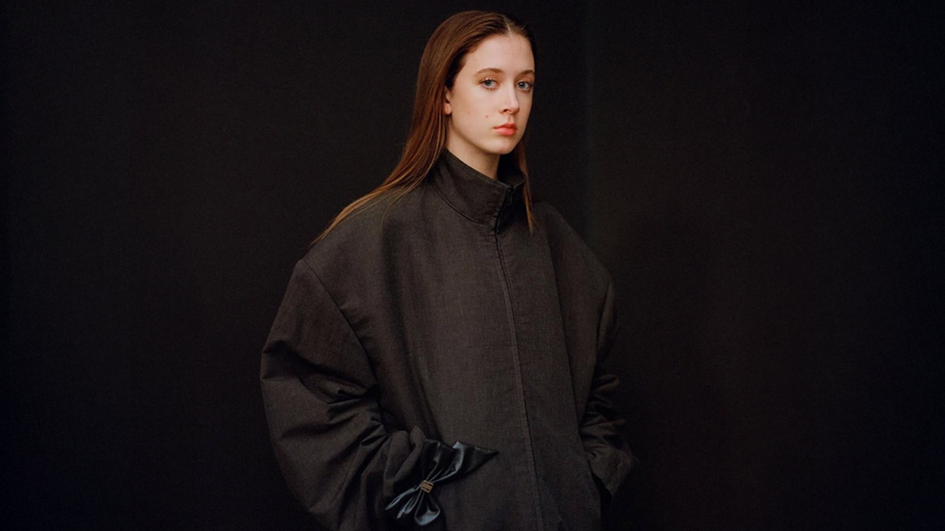 Sunday Rose Kidman Urban poses in a high-fashion editorial photo, wearing an oversized dark jacket with her hands in the pockets, against a black background.