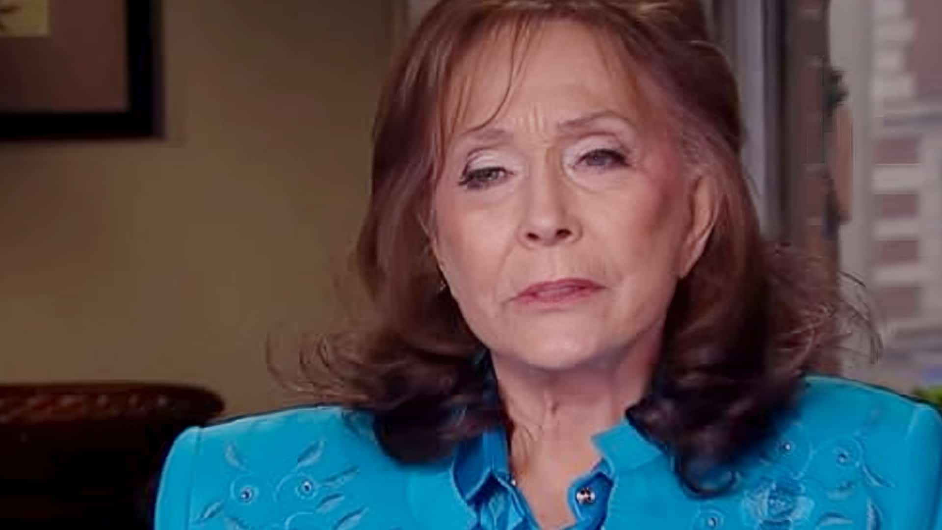 Loretta Lynn wears a bright blue embroidered blouse during a candid interview, her expression serious and heartfelt.