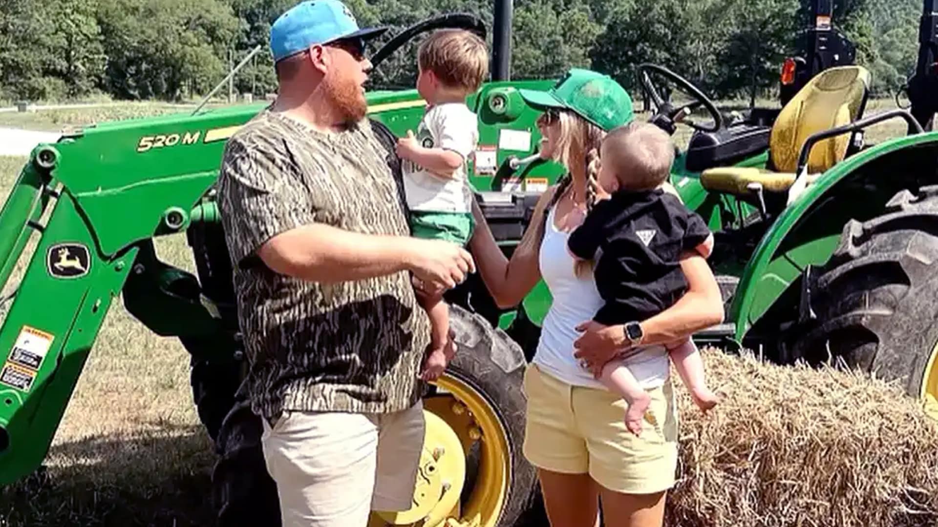 Luke Combs stands with his wife and two sons in front of a green tractor, embracing simple farm life in casual summer clothes.