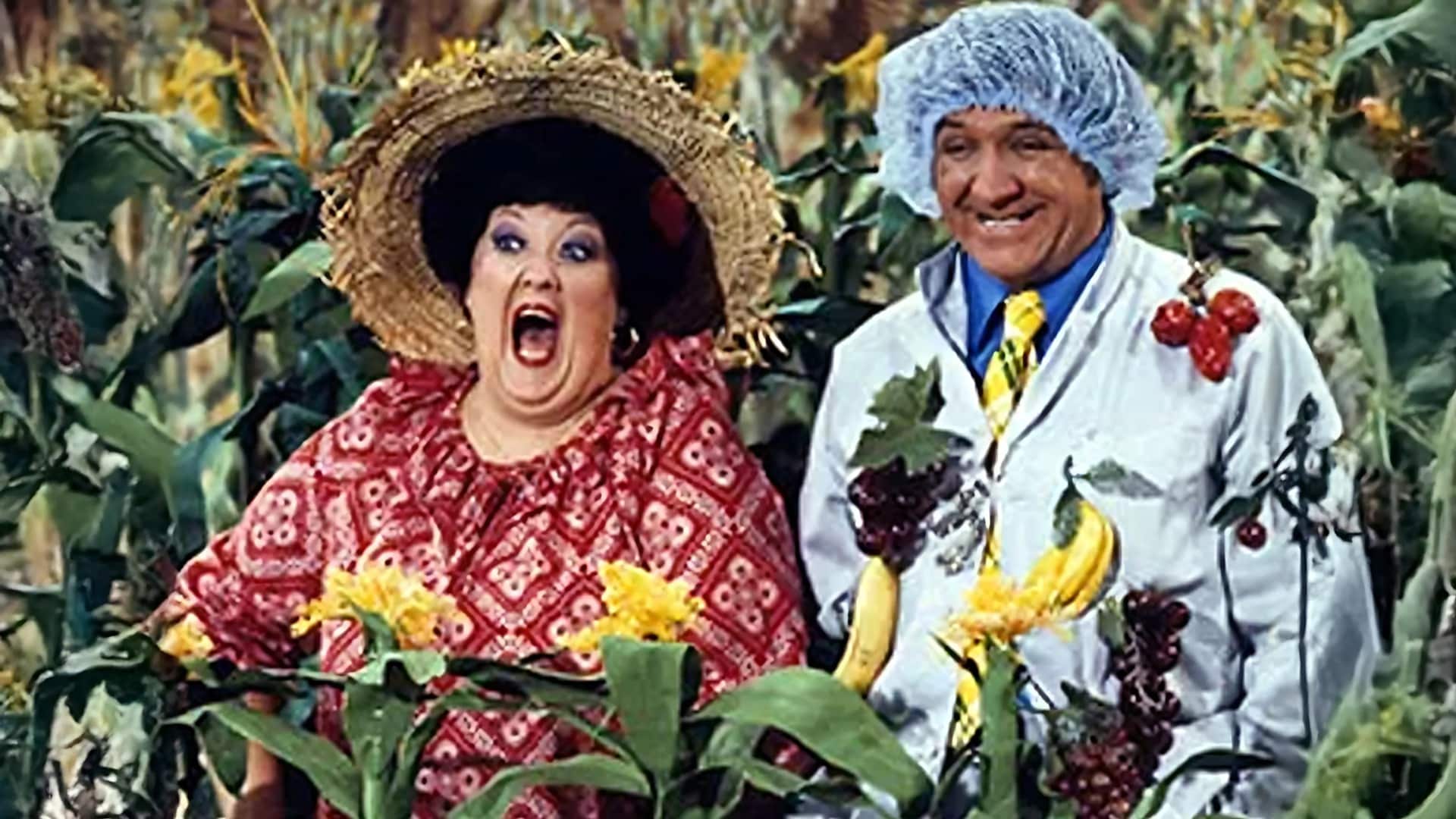 Lulu Roman, dressed in her signature red dress and straw hat, shares a joyful moment on the Hee Haw set—a snapshot of the comedic brilliance and heart that made her a television icon and gospel favorite, now remembered fondly following her passing at 78.