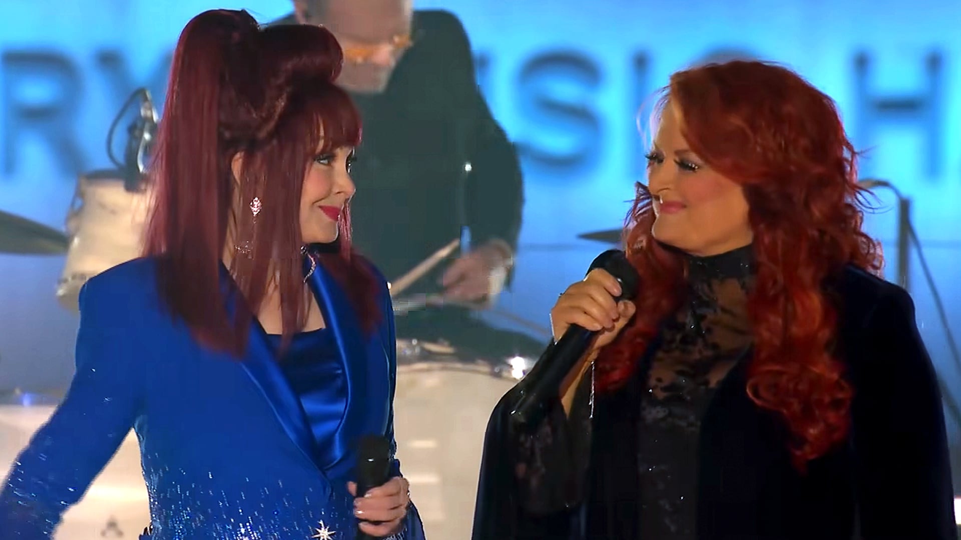 Naomi Judd and Wynonna Judd share a heartfelt look while holding microphones during their final performance, dressed in vibrant stage outfits under concert lighting.