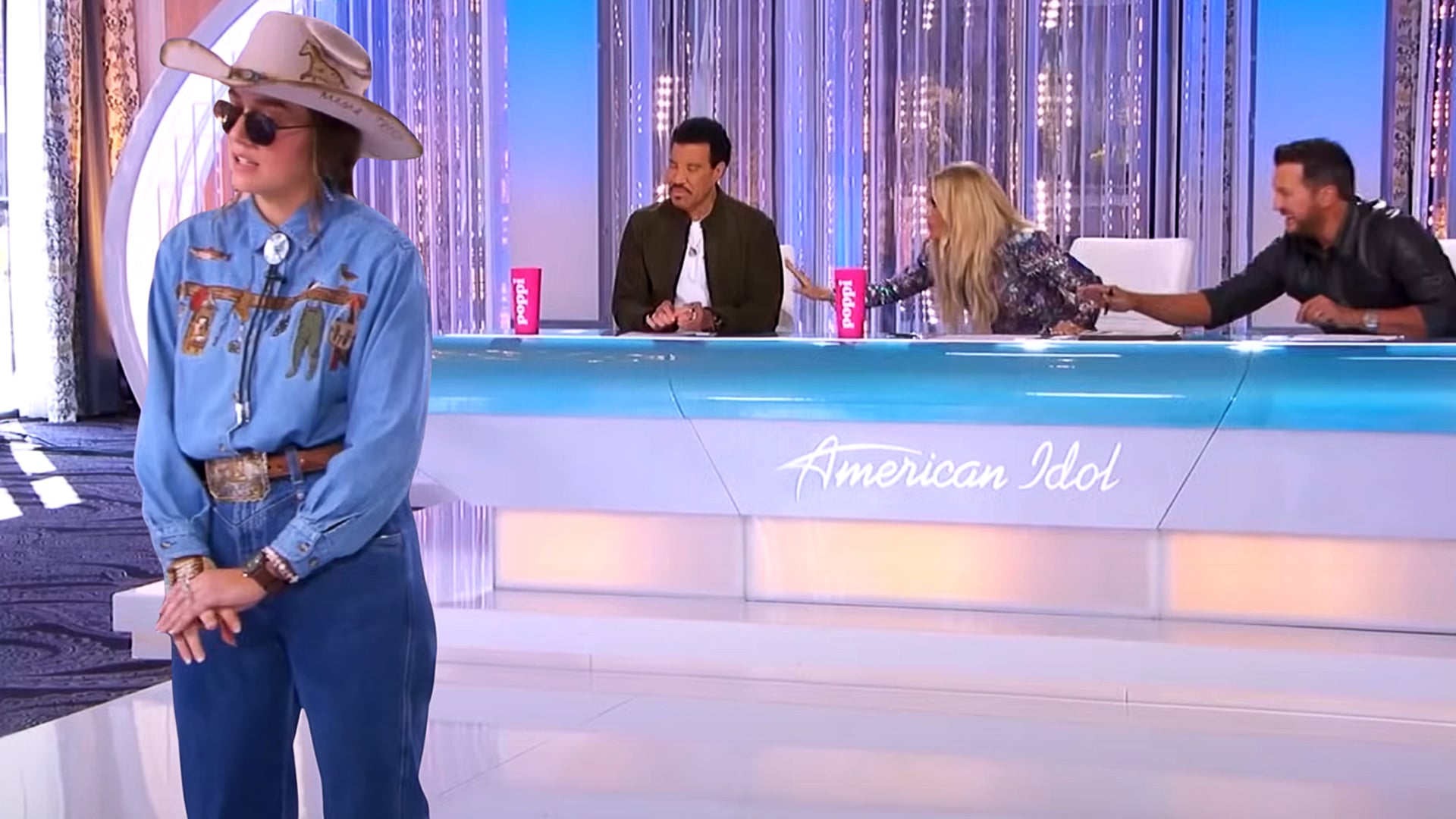 Abi Bowen, wearing a cowboy hat and denim outfit, sings with her back partially turned to the American Idol judges as Carrie Underwood reacts emotionally, reaching across the table.