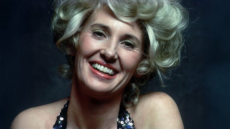 Tammy Wynette smiles warmly in a close-up portrait wearing a sequined black halter top, her platinum blonde hair styled in voluminous curls against a dark background.