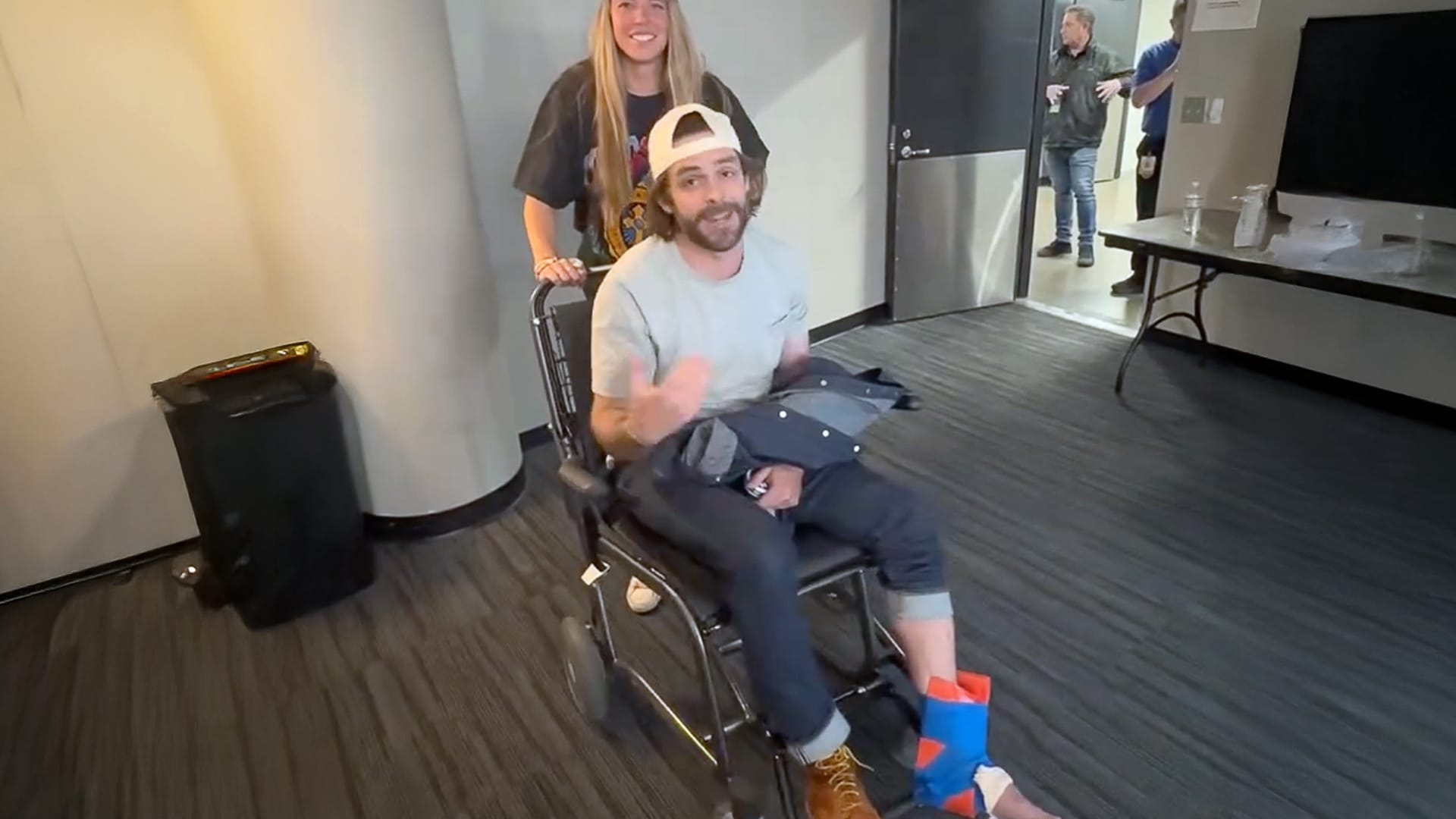 Thomas Rhett sits in a wheelchair with a wrapped ankle, smiling and waving as his wife, Lauren, pushes him backstage. The couple laughs together after his onstage injury during a surprise concert entrance.