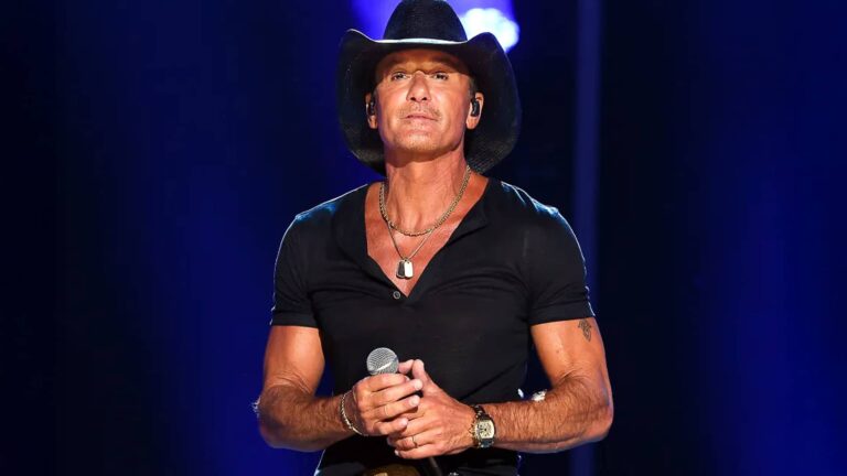 Tim McGraw stands onstage holding a microphone, wearing a black cowboy hat and dark shirt, ready for his 2025 comeback after recovering from multiple surgeries.