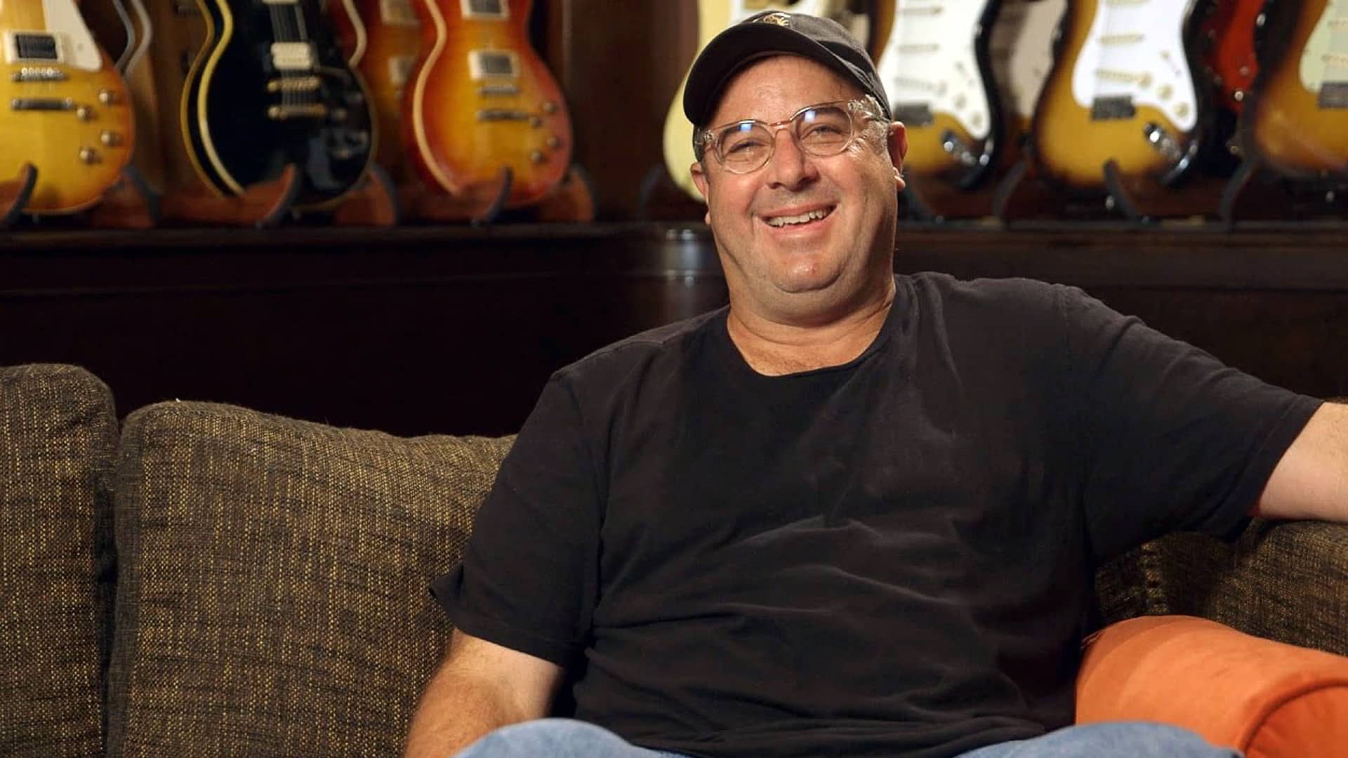 Vince Gill sits comfortably on a couch in front of a wall of guitars, smiling warmly during an interview, dressed casually in a black t-shirt and baseball cap.