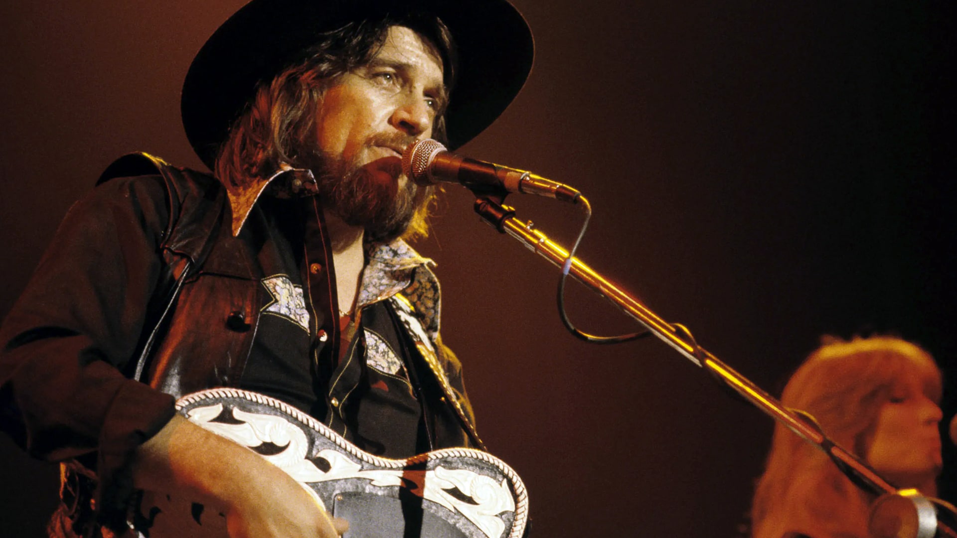 Waylon Jennings performs onstage wearing a black cowboy hat and leather vest, singing into a microphone with a serious expression.