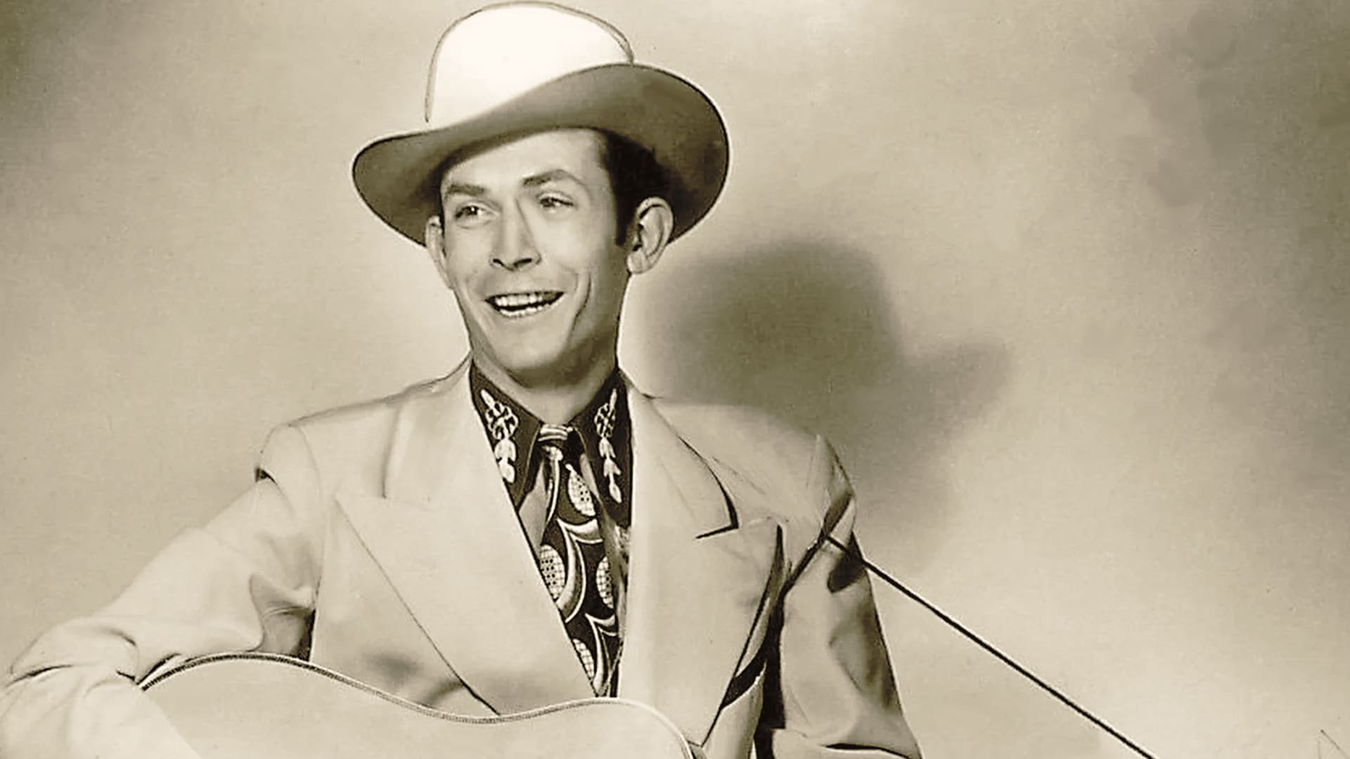 Hank Williams Sr. smiles in a vintage portrait with his guitar, a haunting reminder of the country icon we lost too soon—and the legacy that could've shaped decades more of real country music.