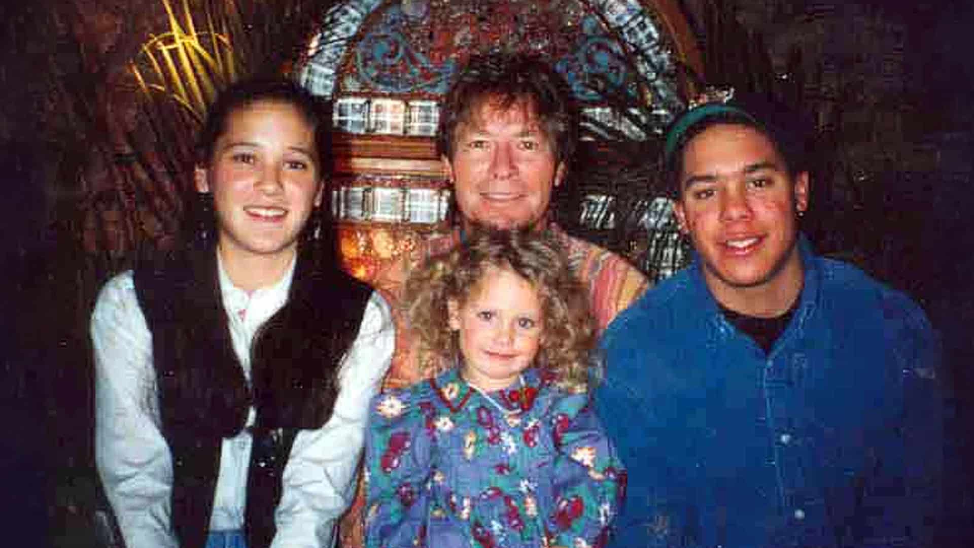 John Denver smiles alongside his three children—Zachary, Anna Kate, and Jesse Belle—in a cherished family photo, representing the legacy they continue to carry forward today.
