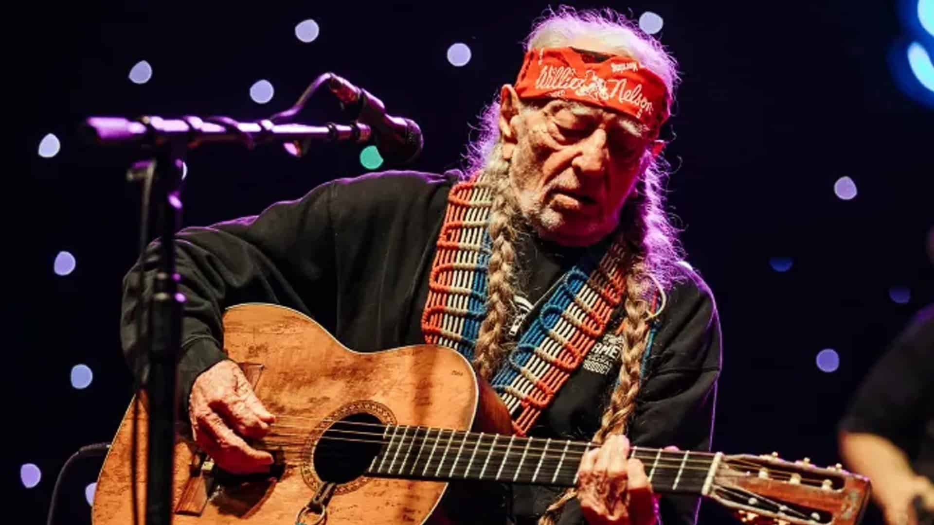 Willie Nelson, wearing a red bandana and black sweatshirt, plays his iconic guitar on stage at 91, bathed in soft stage lights.