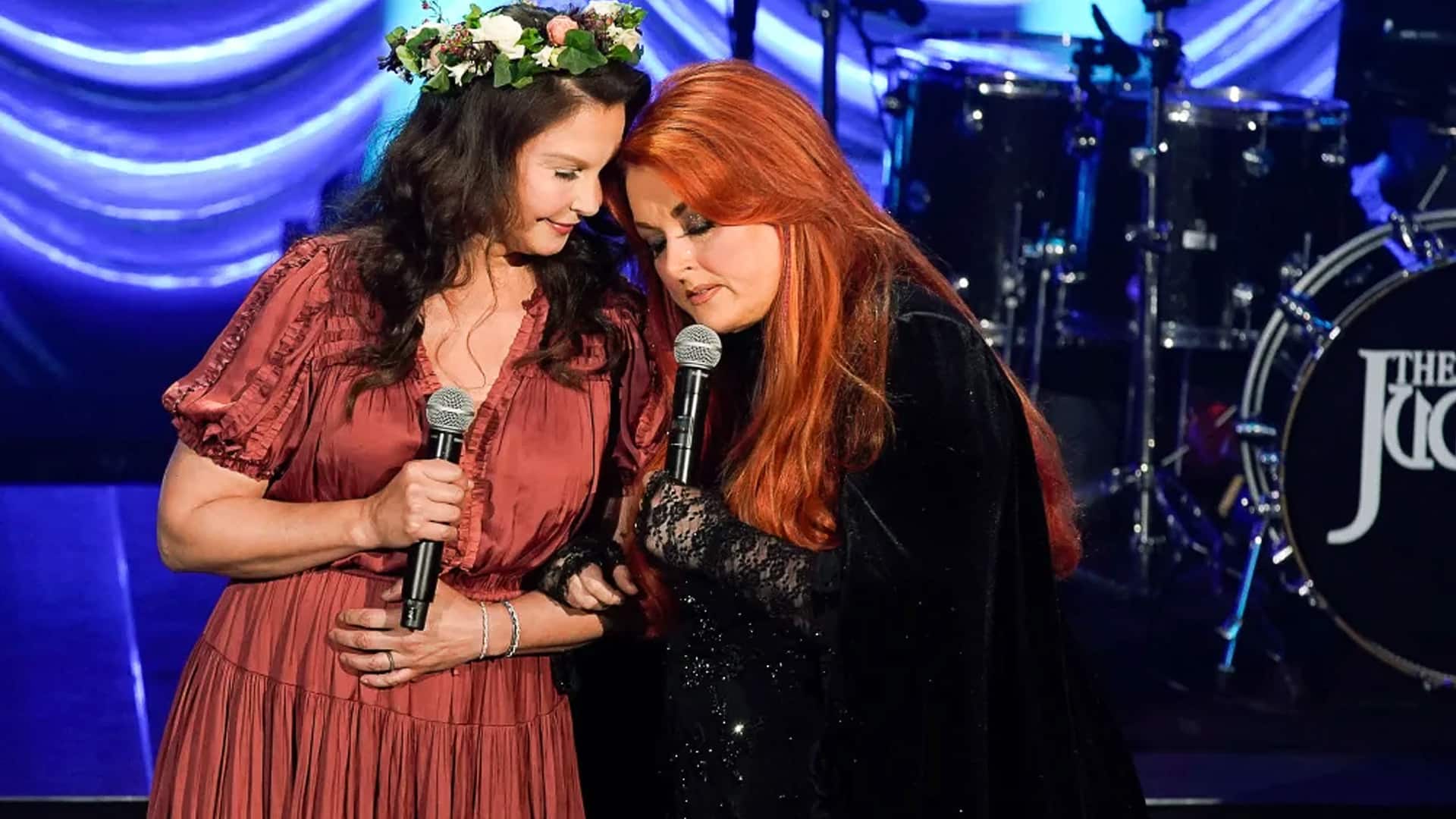 Wynonna Judd and Ashley Judd share an emotional moment onstage, leaning together while holding microphones, with a drum set behind them labeled "The Judds."