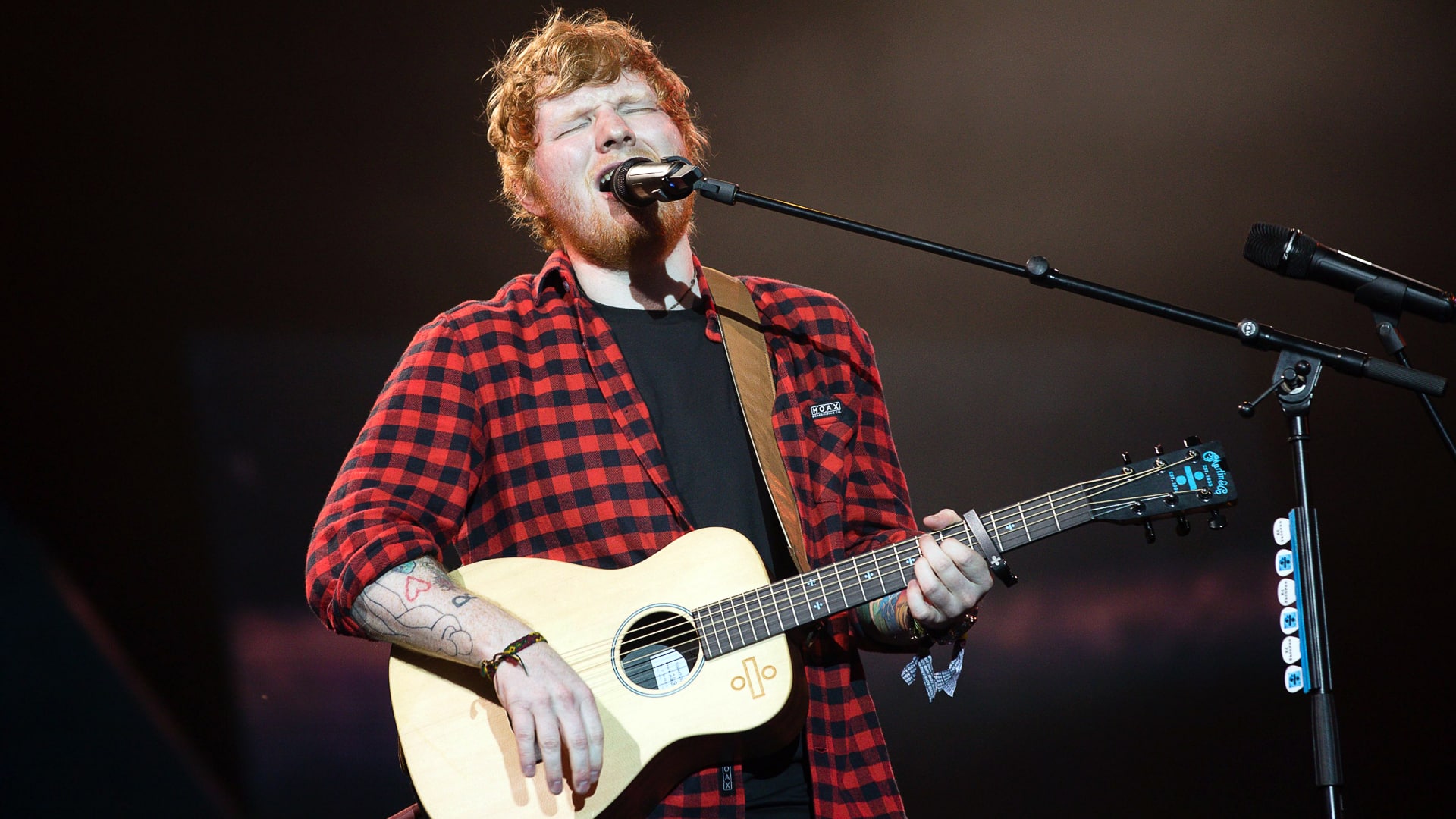 Ed Sheeran performing live in a red flannel shirt, eyes closed mid-chorus, strumming his acoustic guitar with raw emotion and zero twang fakery.