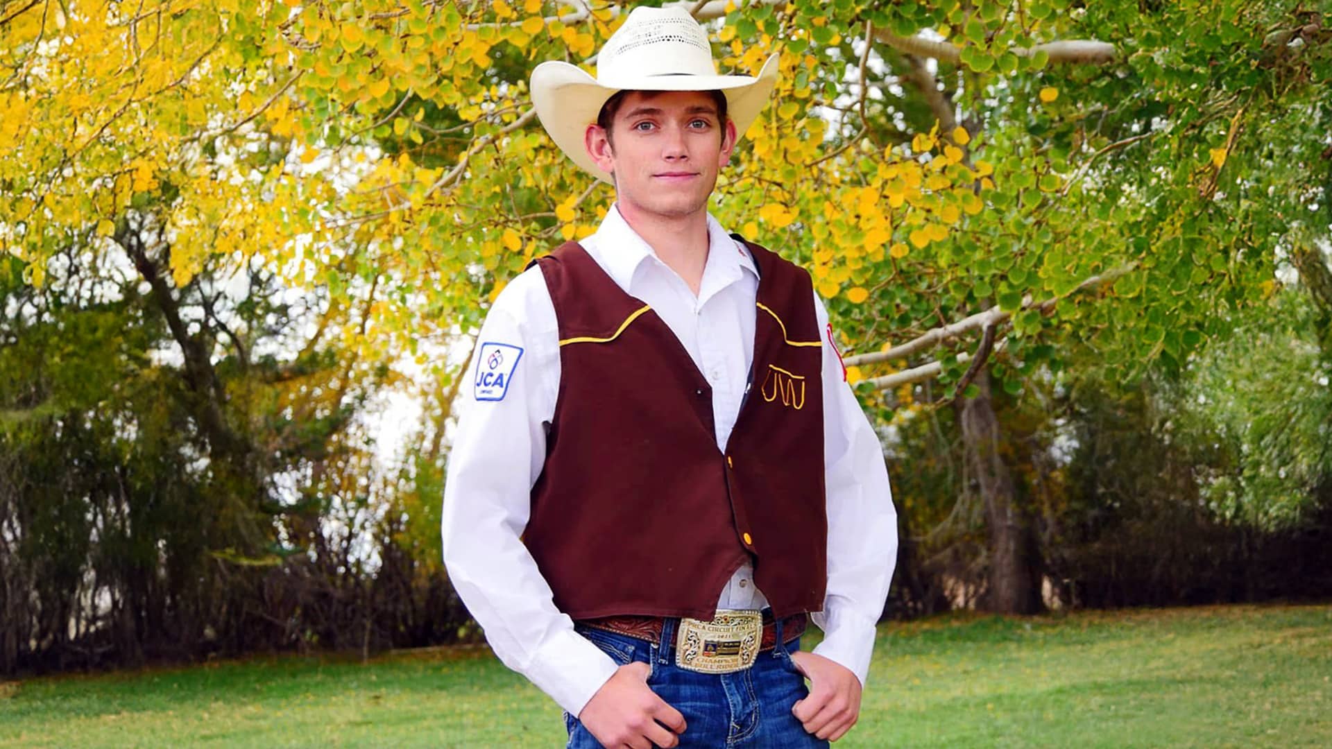 Young bull rider Dylan Grant, dressed in a cowboy hat, white shirt, brown rodeo vest, and championship belt buckle, stands outdoors in front of autumn trees with green and yellow leaves.