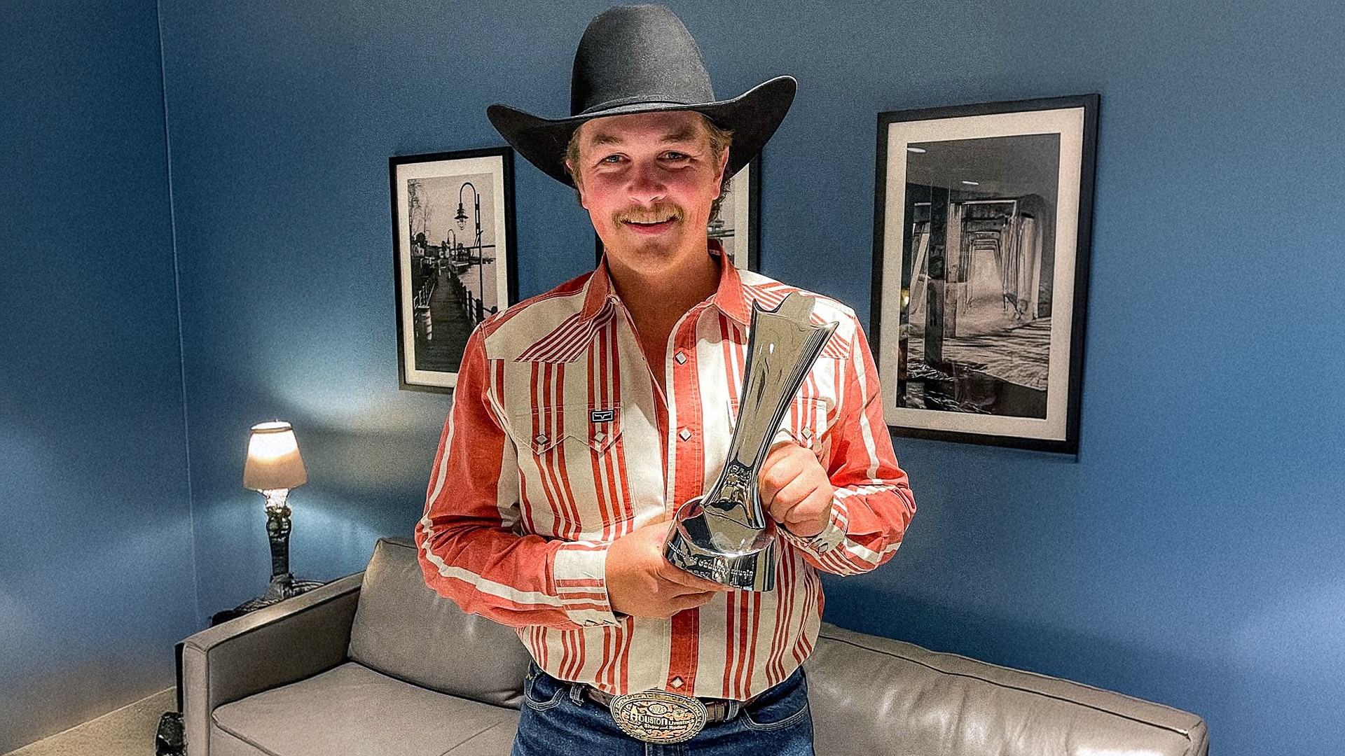 Zach Top smiles while proudly holding his ACM New Male Artist of the Year trophy, dressed in a red-striped western shirt and black cowboy hat.