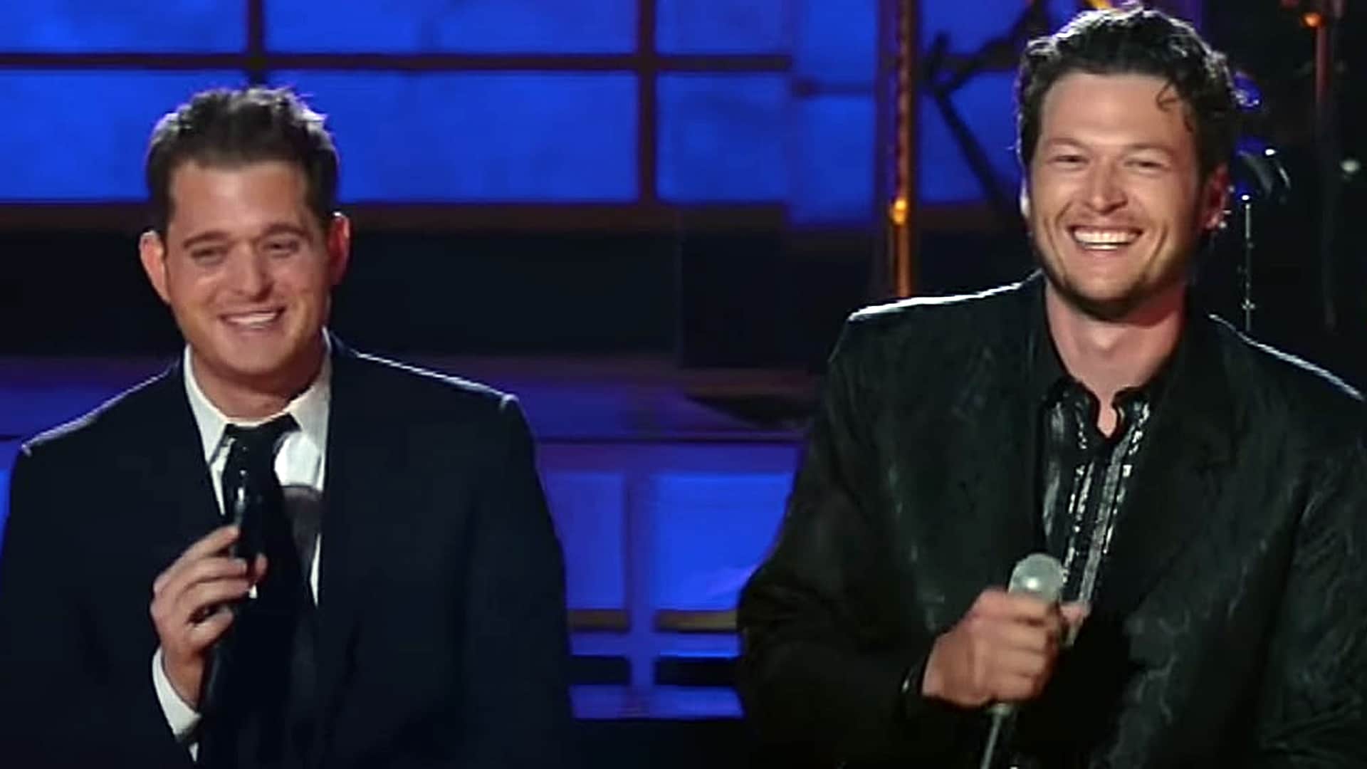 Blake Shelton and Michael Bublé smile onstage while holding microphones during their performance, blending country and pop charm under bright stage lights.