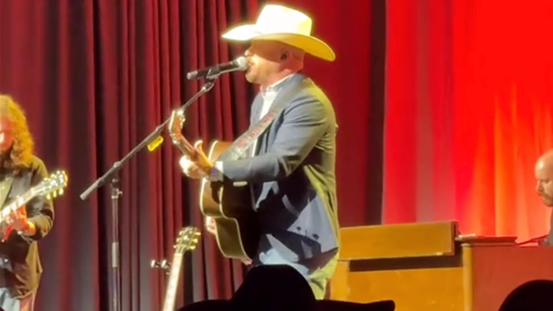 Cody Johnson performs a heartfelt rendition of “Red Dirt Road” in a cowboy hat and suit jacket at the ACM Gala, backed by a warm red stage curtain and live band.