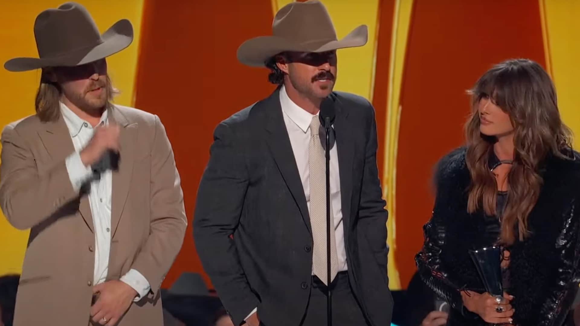 Ella Langley and Riley Green stand onstage at the 2025 ACM Awards accepting the Single of the Year trophy for “You Look Like You Love Me,” flanked by their team and lit by golden stage lights.