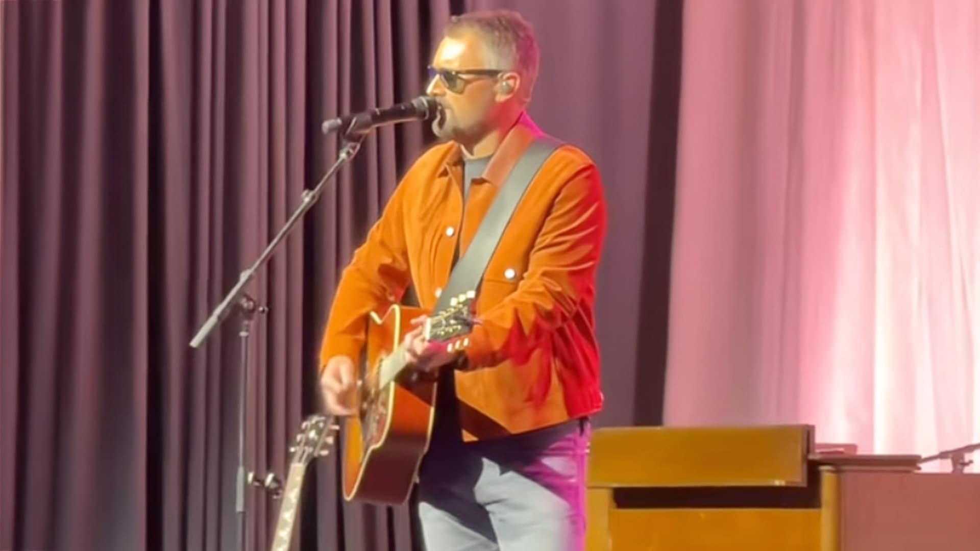 Eric Church performs an acoustic tribute in an orange jacket and sunglasses, playing guitar under warm stage lighting during the ACM gala in honor of Brooks & Dunn.