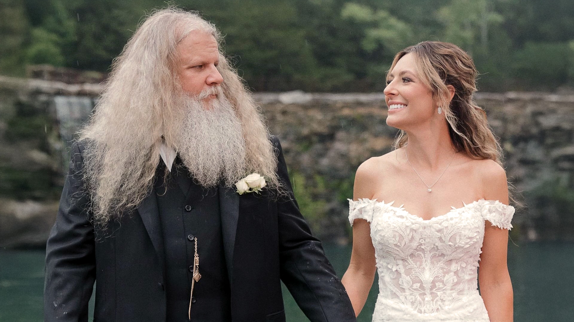 Jamey Johnson and his wife Brittney Eakins smile at each other in wedding attire by a scenic river, celebrating their unique love story that began with a proposal the day after they met.