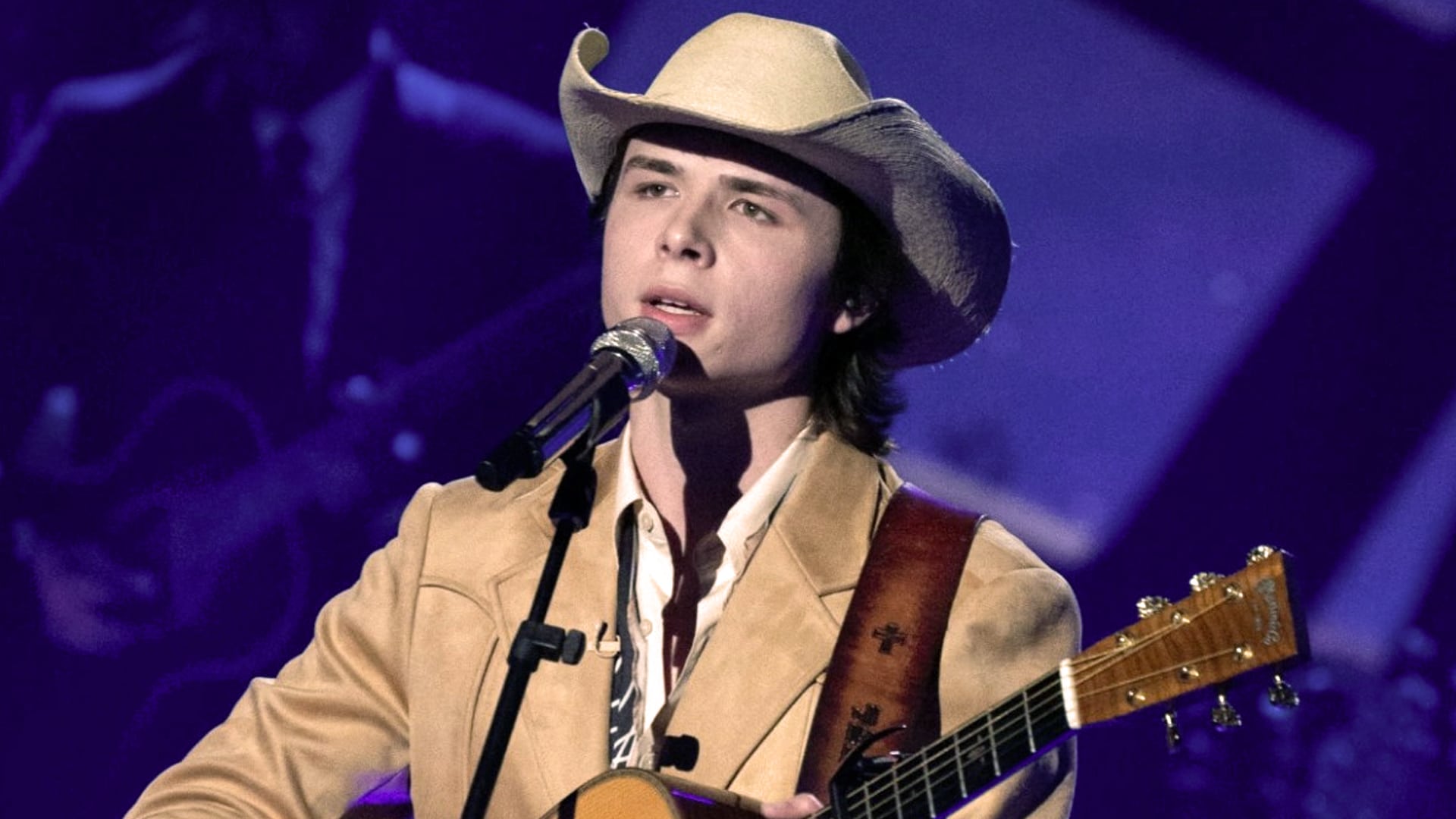 John Foster performs onstage in a tan jacket and cowboy hat, holding a guitar and singing into a microphone under purple lighting.