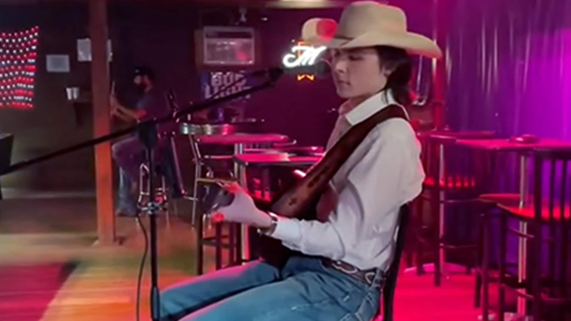 John Foster sits onstage in a dimly lit honky-tonk, performing a heartfelt acoustic cover of "Amarillo by Morning" in a cowboy hat and jeans.