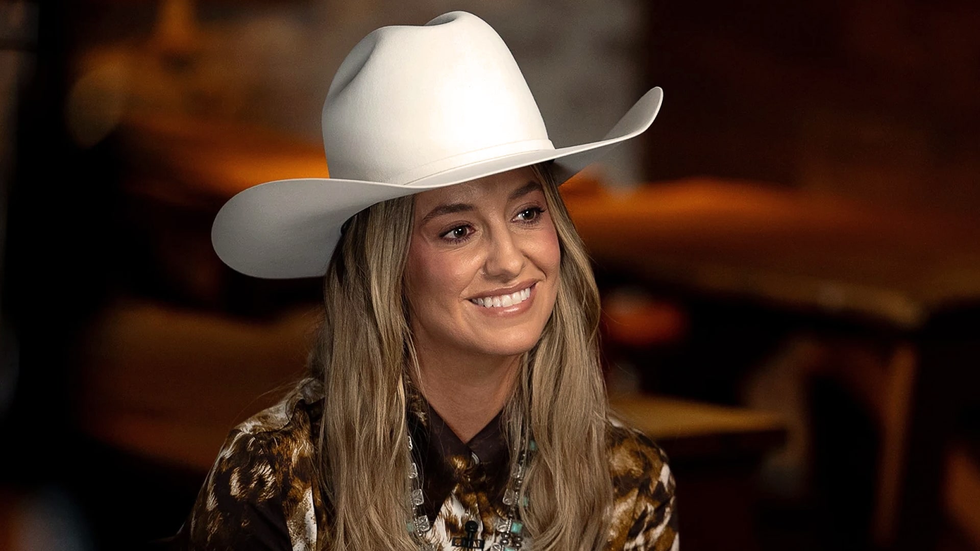 Lainey Wilson smiles in a white cowboy hat, reflecting the grit and journey from her camper days to country music’s biggest stages.