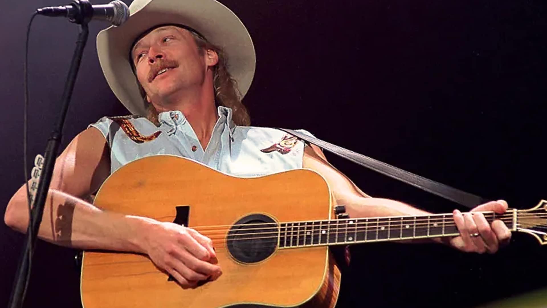 Alan Jackson plays guitar in a sleeveless denim shirt and cowboy hat, embodying his classic country style—the same unapologetic spirit he later showed during his silent protest at the ACM Awards.