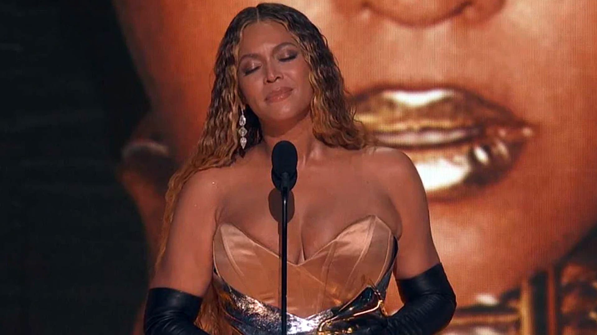 Beyoncé stands at the microphone in a strapless gown and gloves, appearing emotional during an award show—symbolic of the attention she brought to country music with Cowboy Carter, despite being shut out of the 2025 ACM Awards.