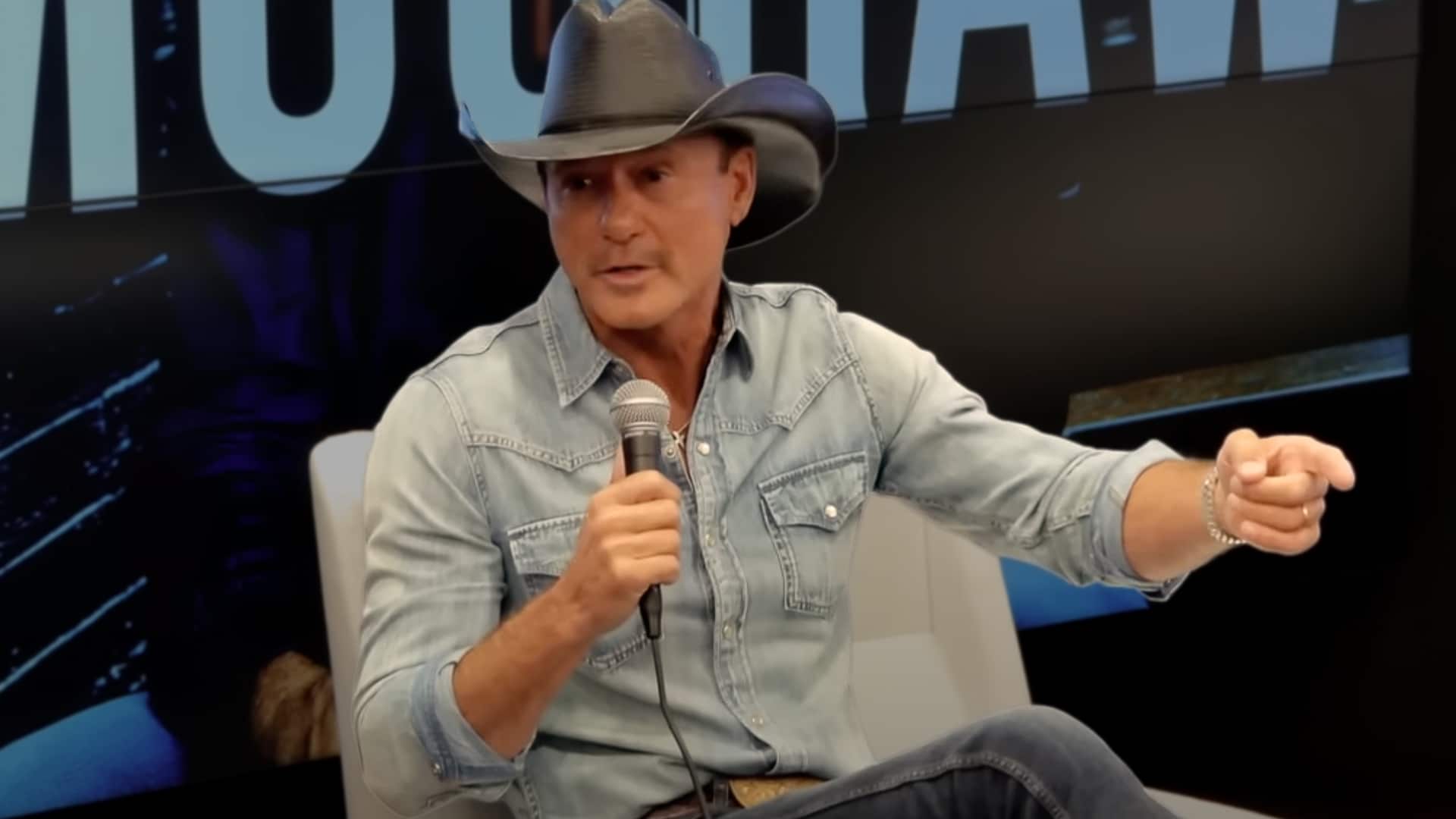 Tim McGraw wears a cowboy hat and denim shirt while speaking into a microphone during a live interview, addressing his recovery after multiple surgeries.