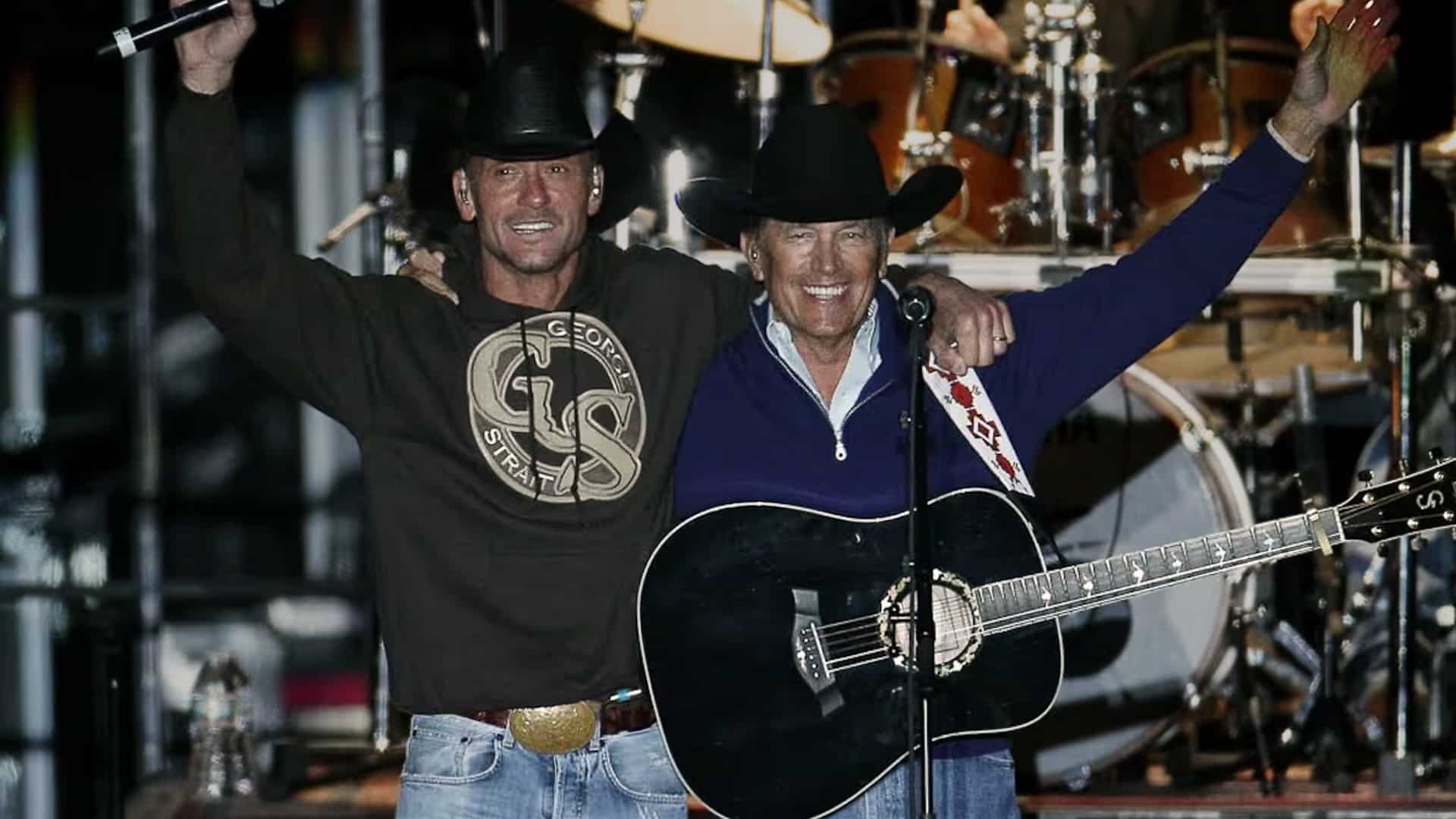 Tim McGraw and George Strait stand side by side onstage, smiling with arms raised and guitars in hand, sharing a moment of camaraderie and mutual respect during a live performance.