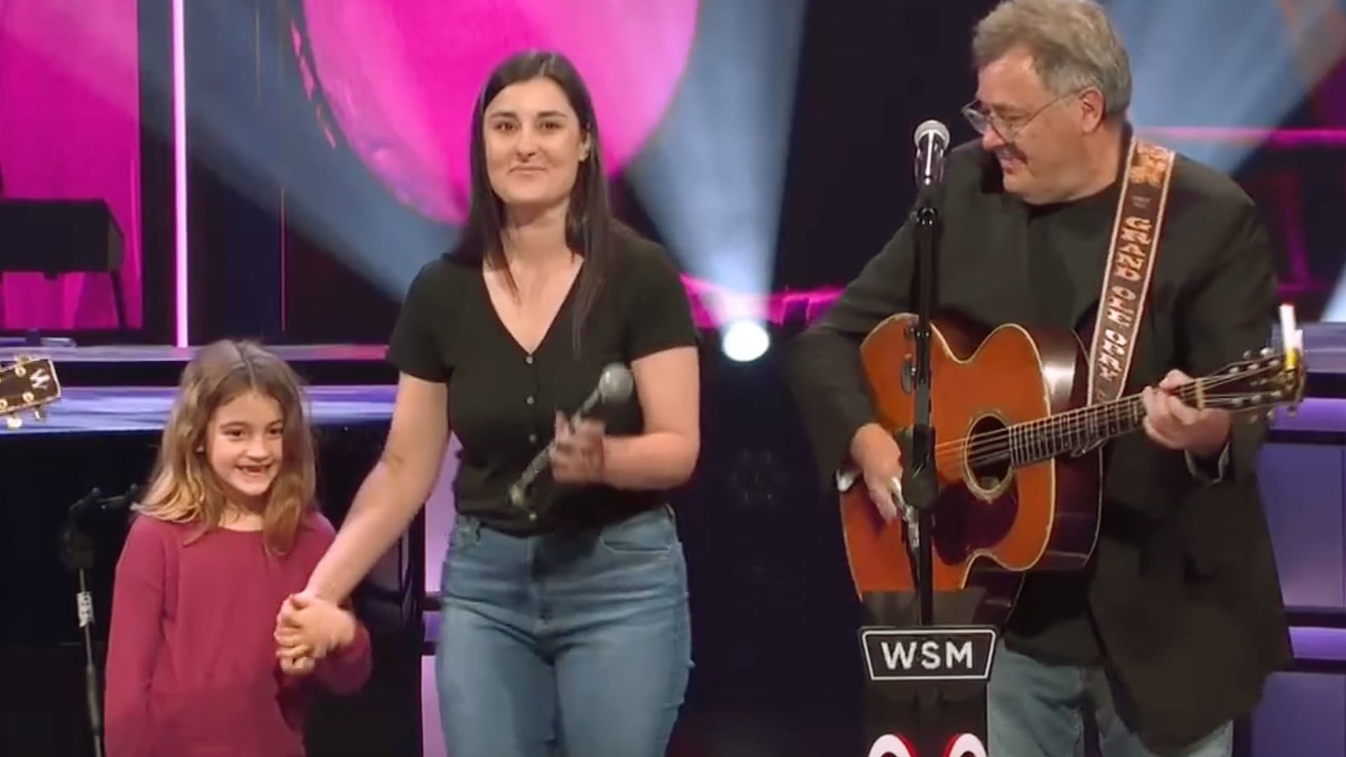 Vince Gill smiles while playing guitar onstage at the Grand Ole Opry as his granddaughter Everly and daughter Jenny Gill join him, creating a touching three-generation family moment.