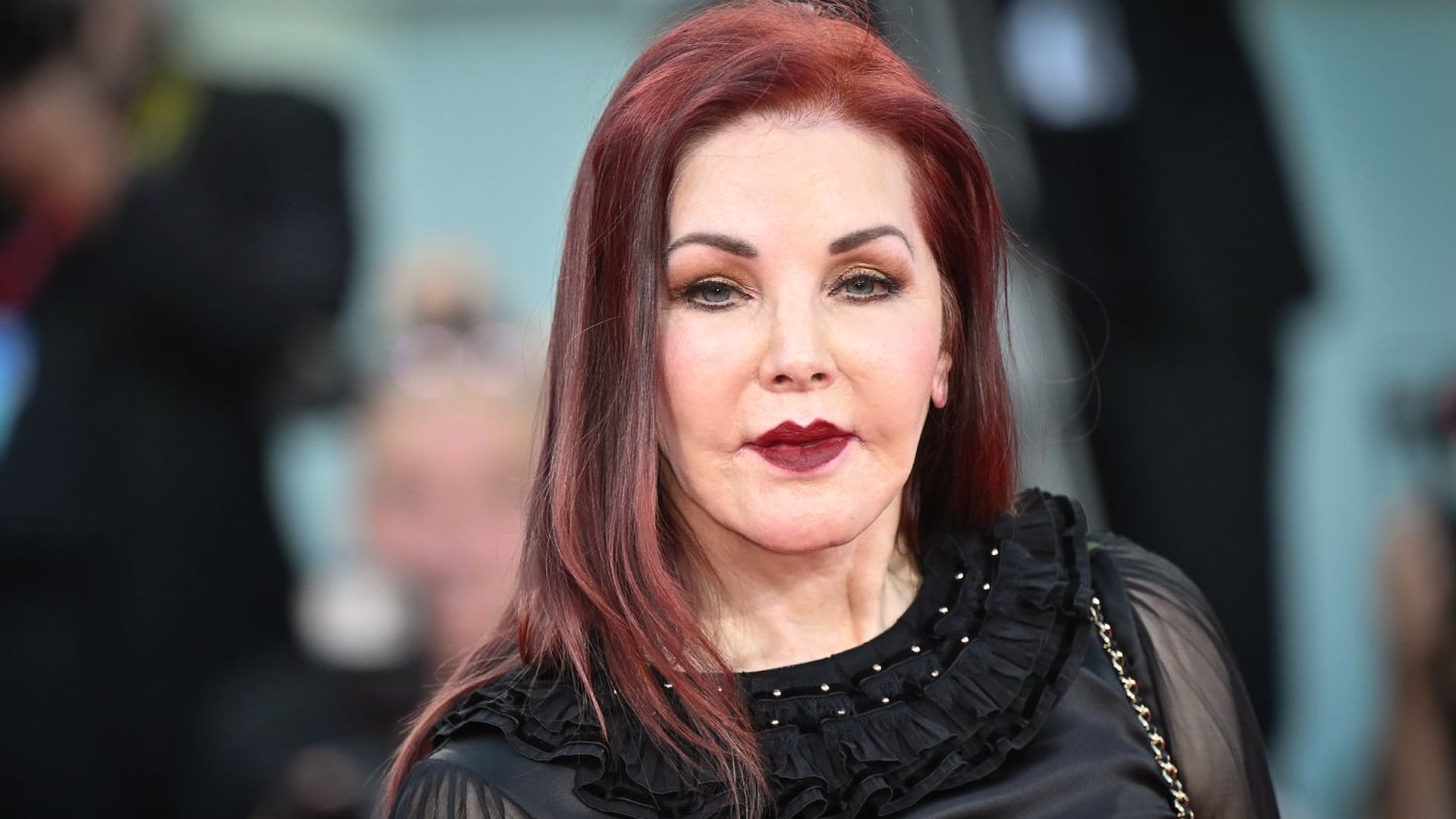 Priscilla Presley poses in a black ruffled blouse with dark red lipstick and auburn hair, attending a formal event years after her life with Elvis Presley, still carrying the legacy of their enduring love story.