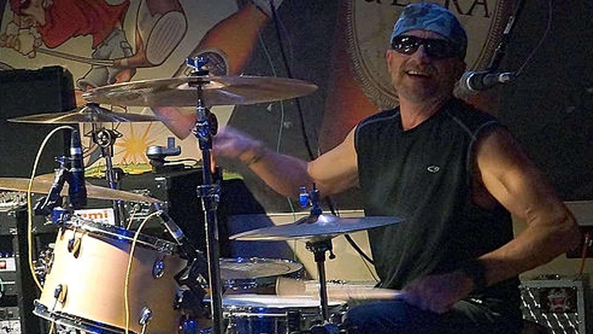 Mark Herndon plays drums on stage with a smile, wearing sunglasses and a bandana, representing his longtime role with country supergroup Alabama despite feeling like an outsider.