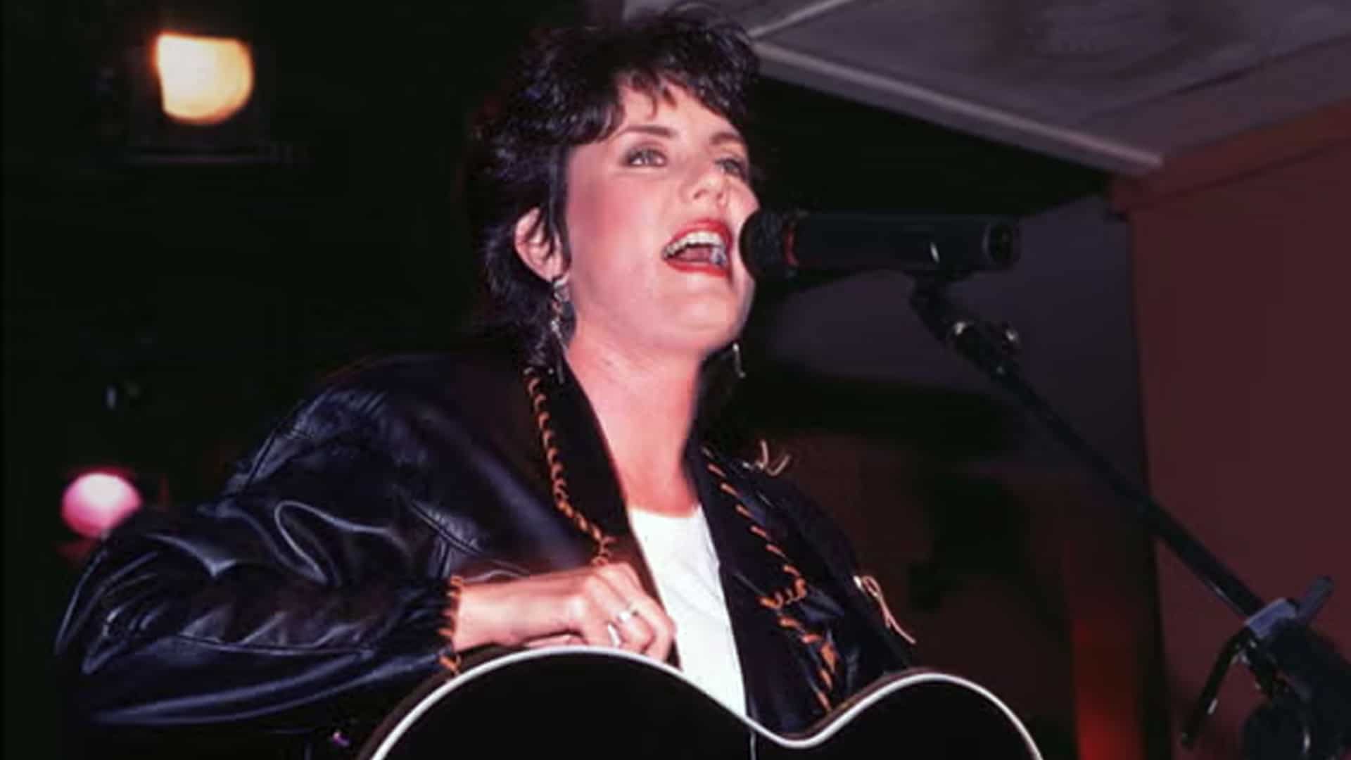 Holly Dunn performs passionately under soft stage lights with her guitar in hand, capturing the emotion behind her heartfelt classic “Daddy’s Hands.”