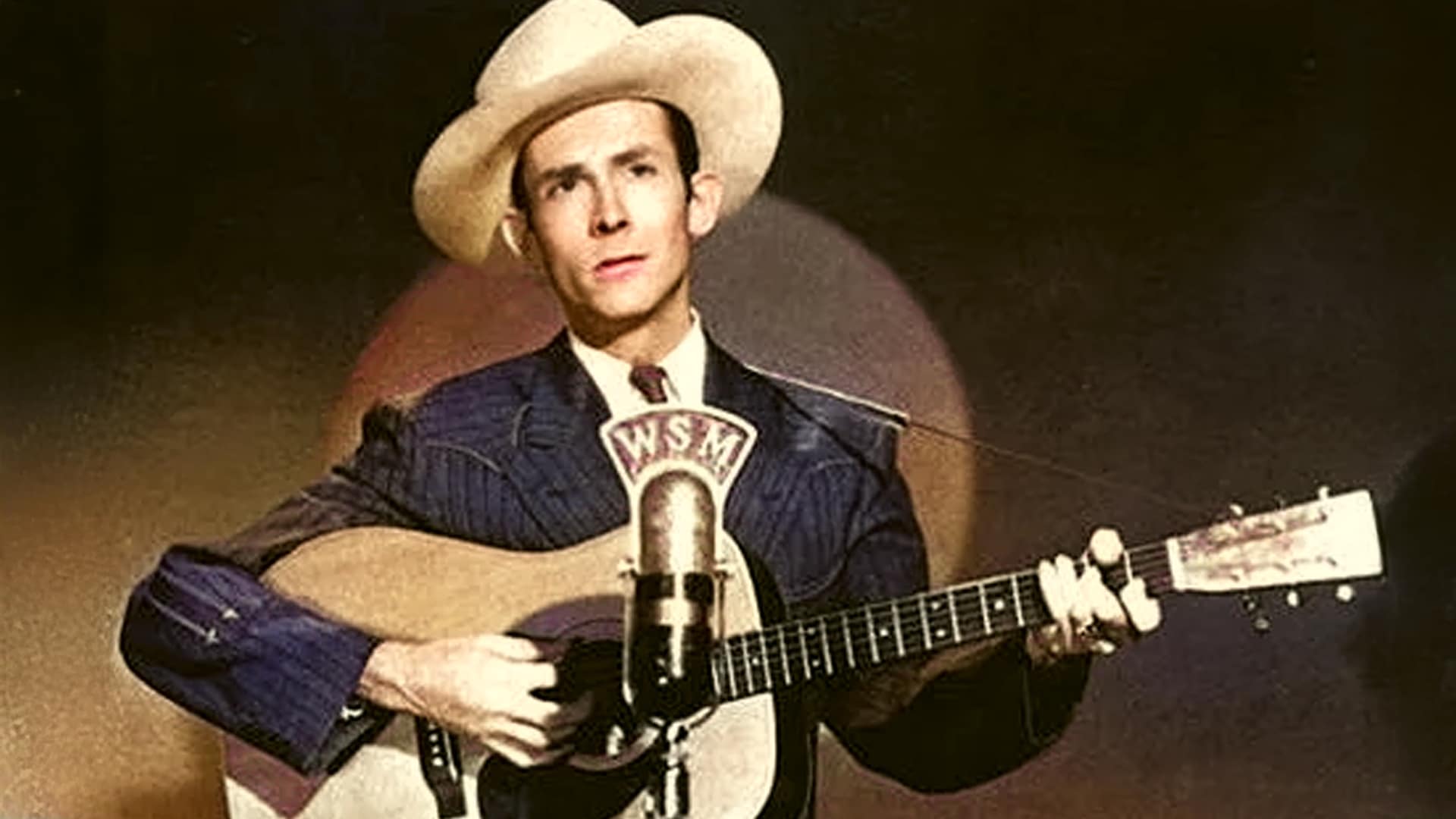 Hank Williams performs “Lovesick Blues” at the Grand Ole Opry in 1949, standing at the WSM microphone in a pinstripe suit and cowboy hat, holding an acoustic guitar during his historic debut.