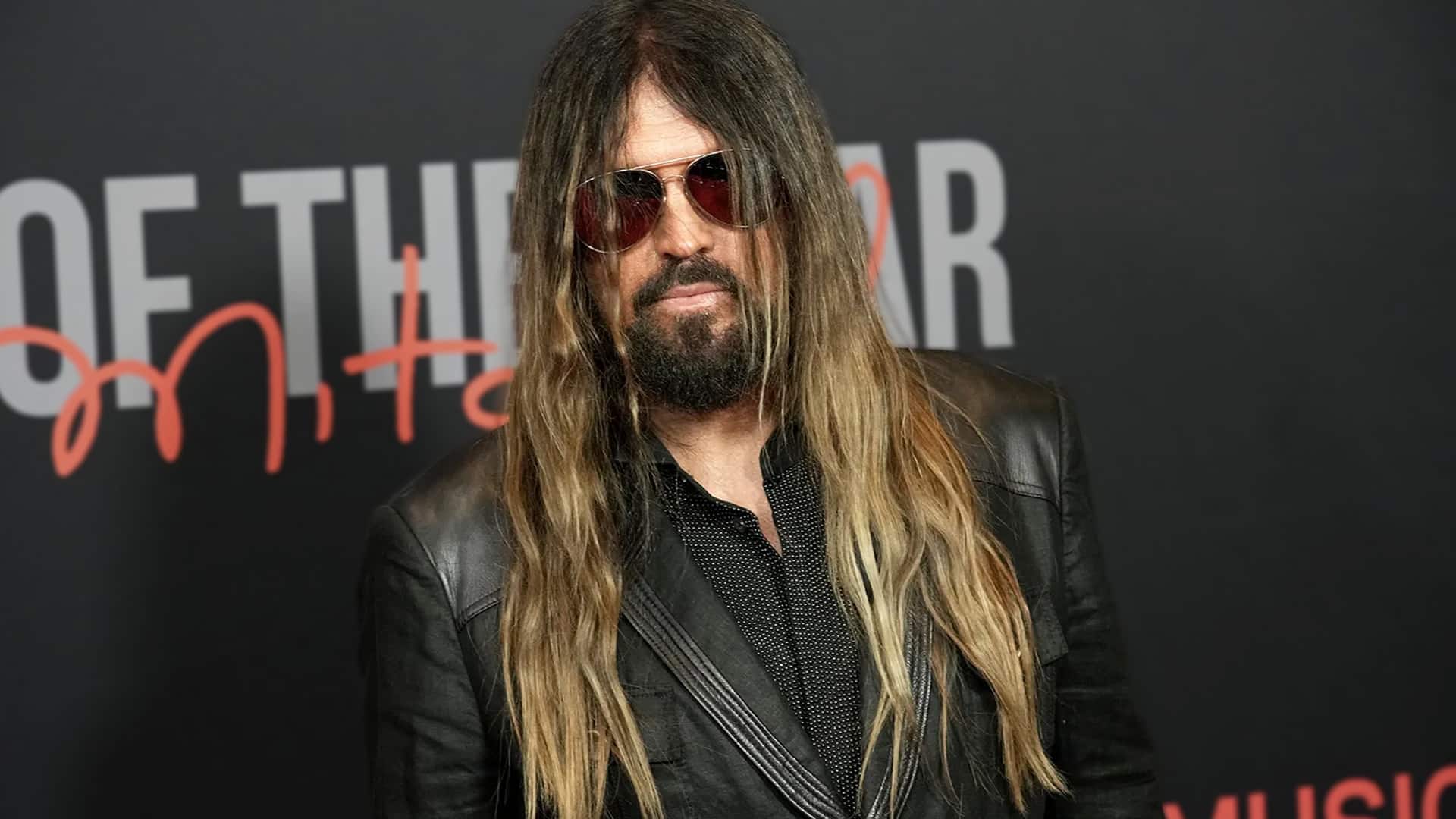 Billy Ray Cyrus stands on the red carpet in a black leather jacket and sunglasses, unintentionally becoming the center of family drama after Trace Cyrus called him out online.