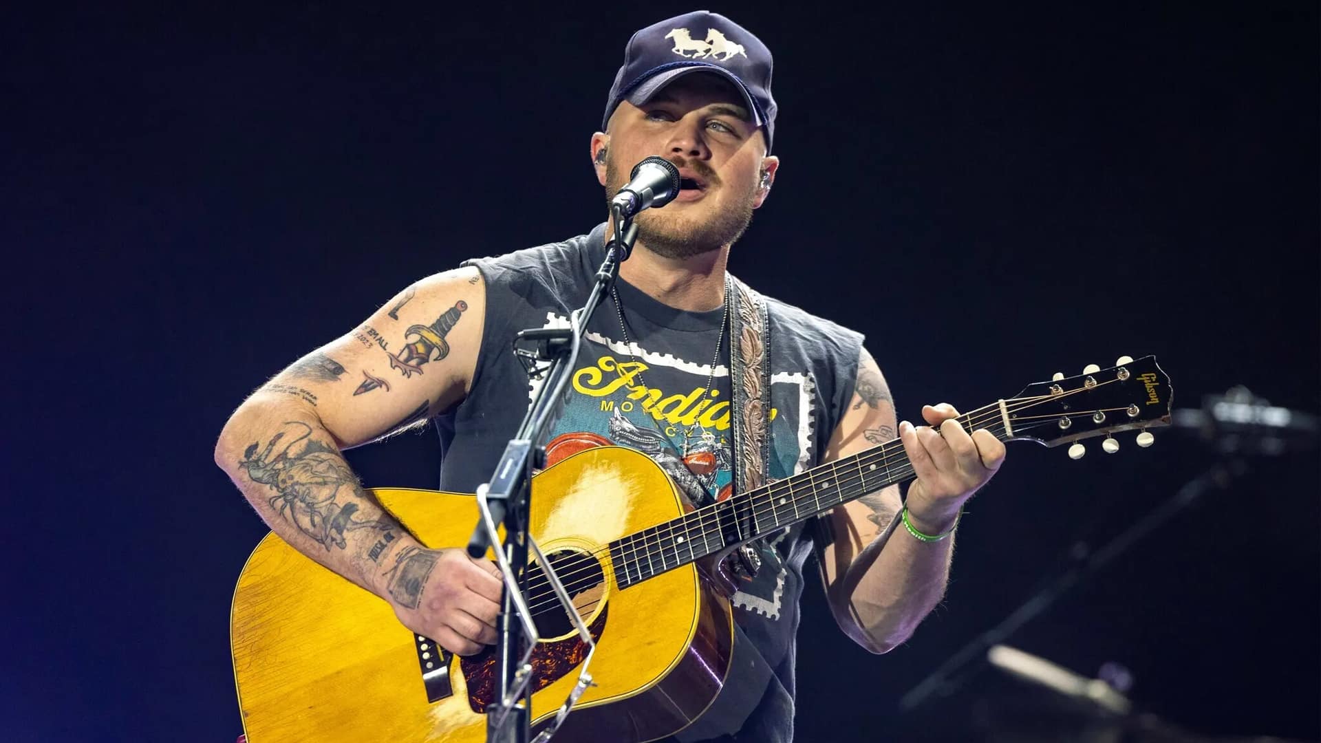 Zach Bryan performs live on stage in a previous concert, wearing a sleeveless shirt and ball cap, as he announces a surprise $50 Red Rocks show set for August.