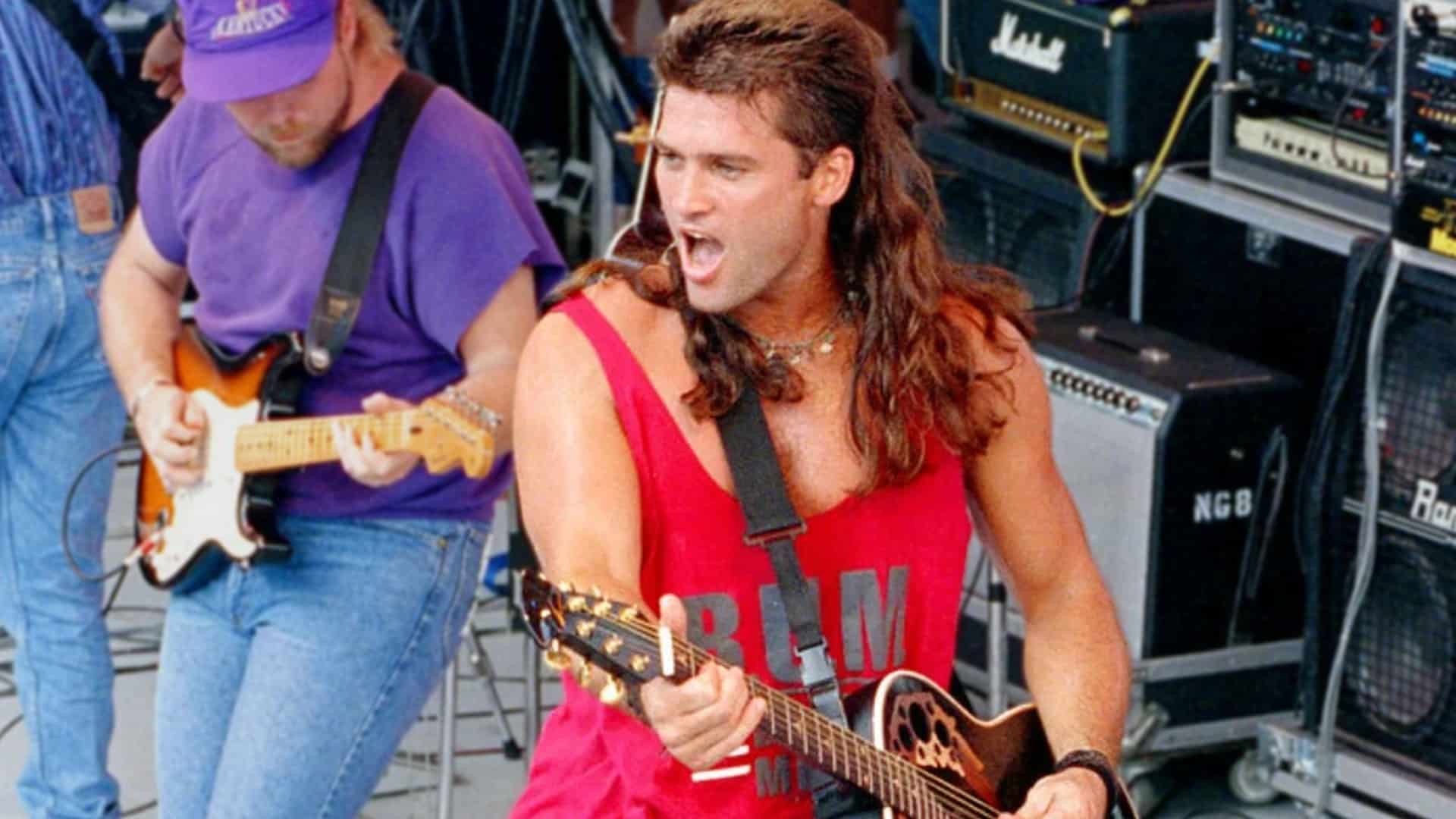 Billy Ray Cyrus performs “Achy Breaky Heart” in 1992 wearing a red tank top and rocking his iconic mullet during a high-energy outdoor concert.