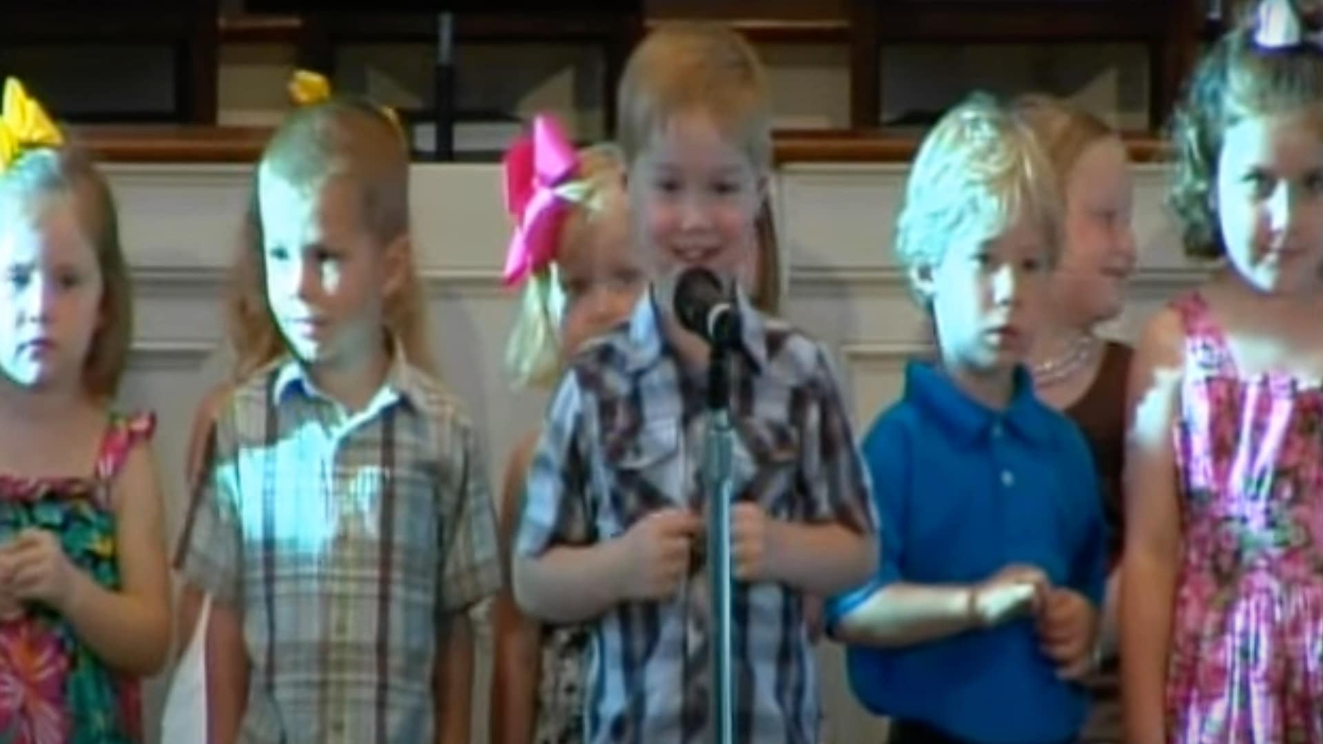 Little Mississippi boy in plaid shirt smiles at mic during preschool graduation where he recited New Testament books and sang a George Strait classic.