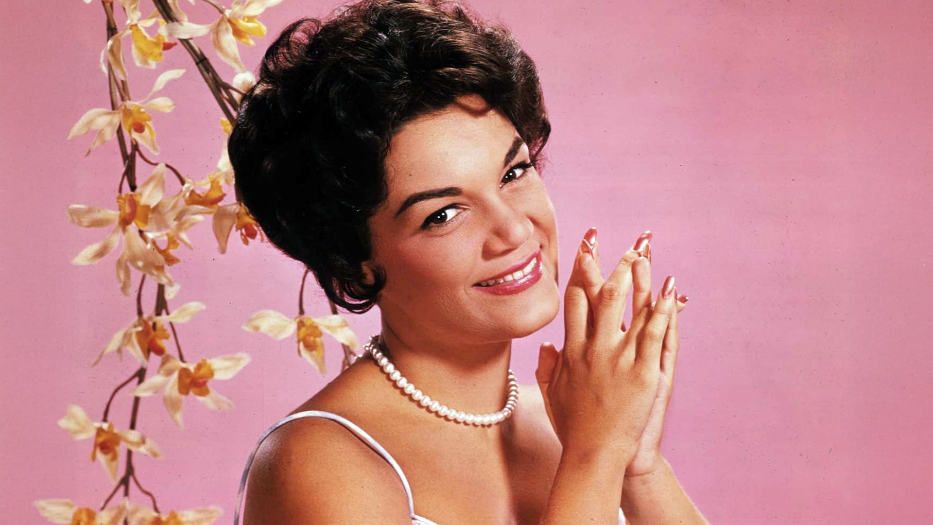 Connie Francis smiles with hands clasped in front of a pink backdrop and orchids, remembered for her timeless hit “Pretty Little Baby.”